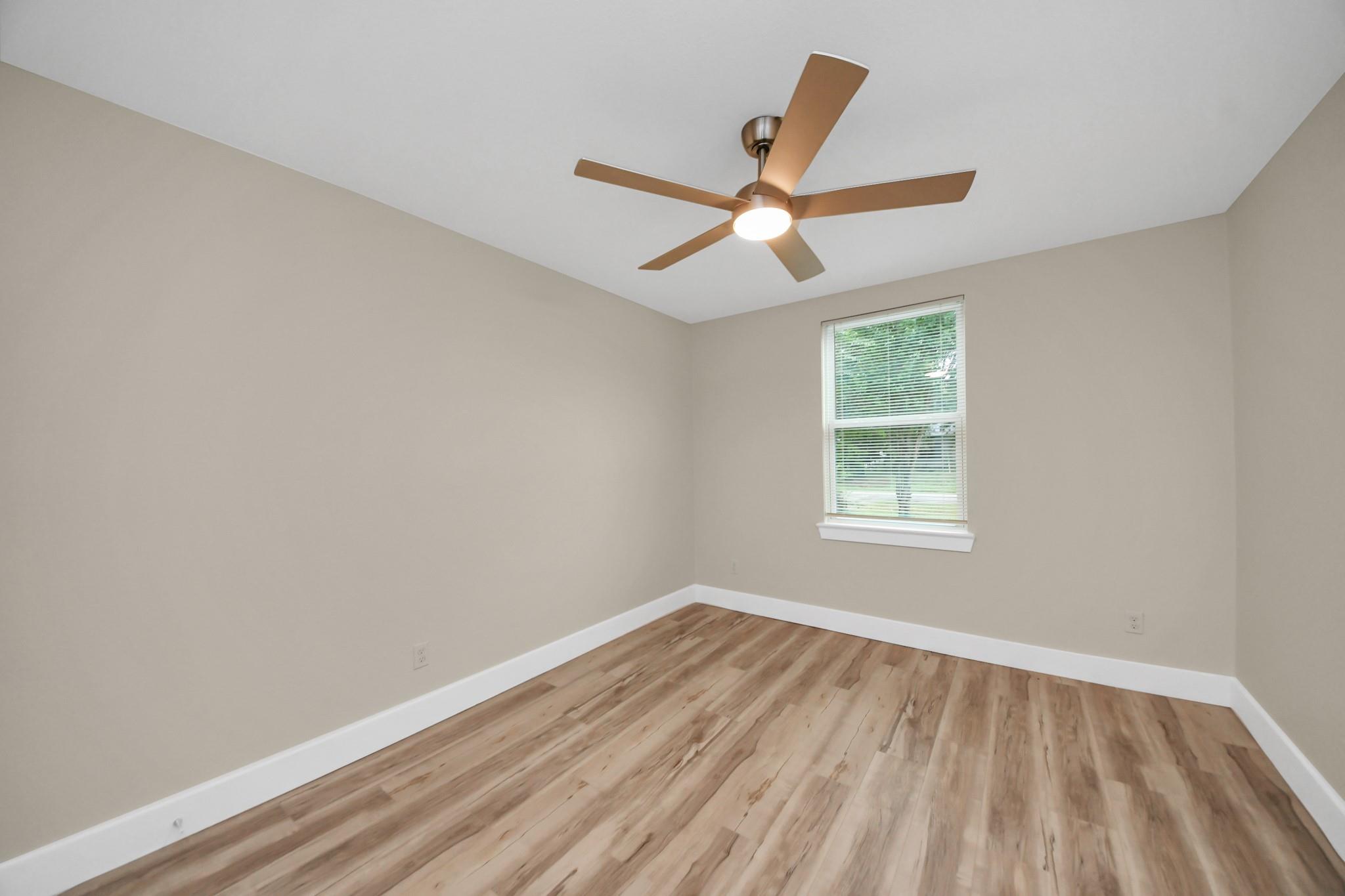 Empty bedroom with wood-look flooring, a window with blinds, and a lit ceiling fan.