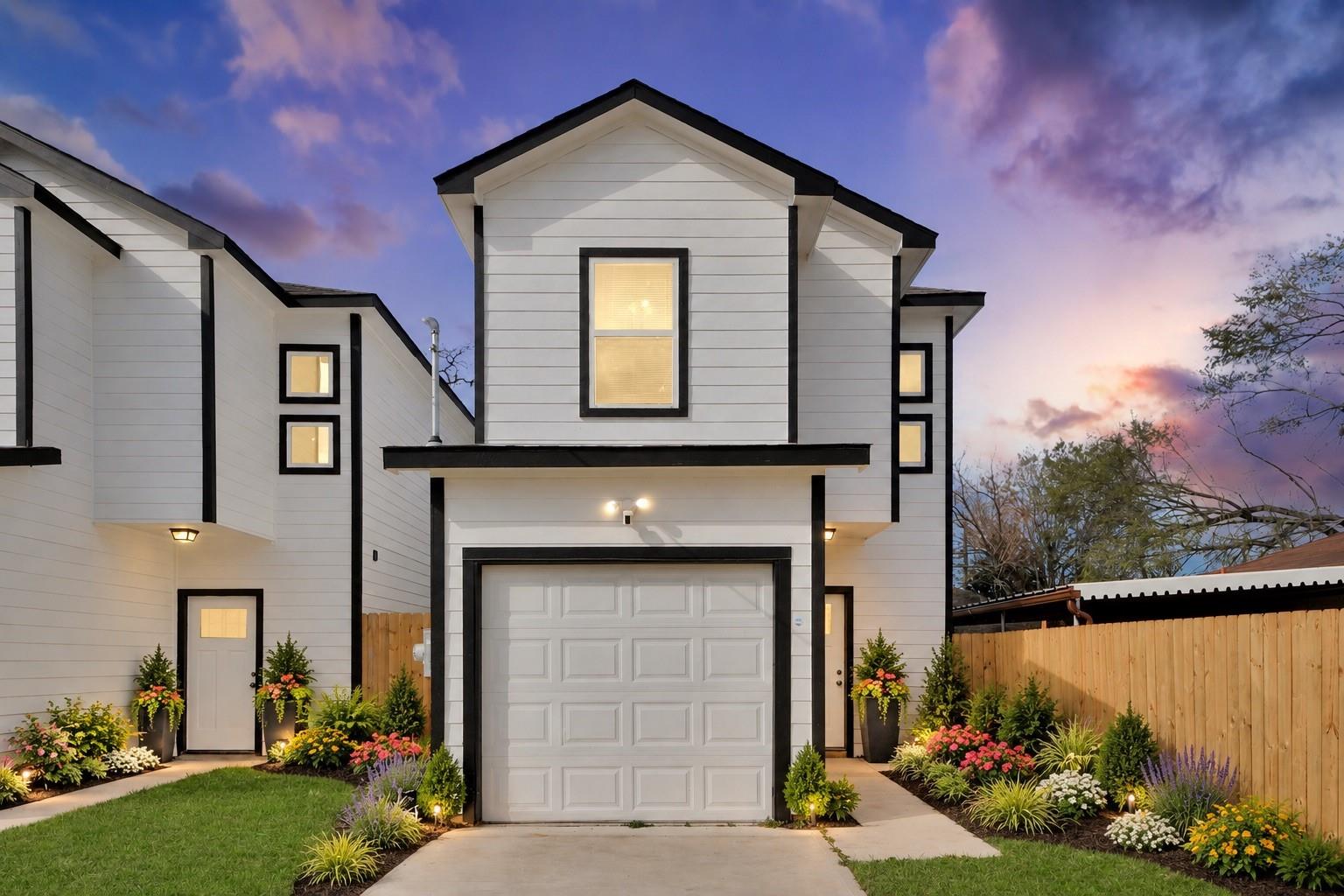 Modern two-story white home with black trim, garage, and colorful landscaping at dusk.