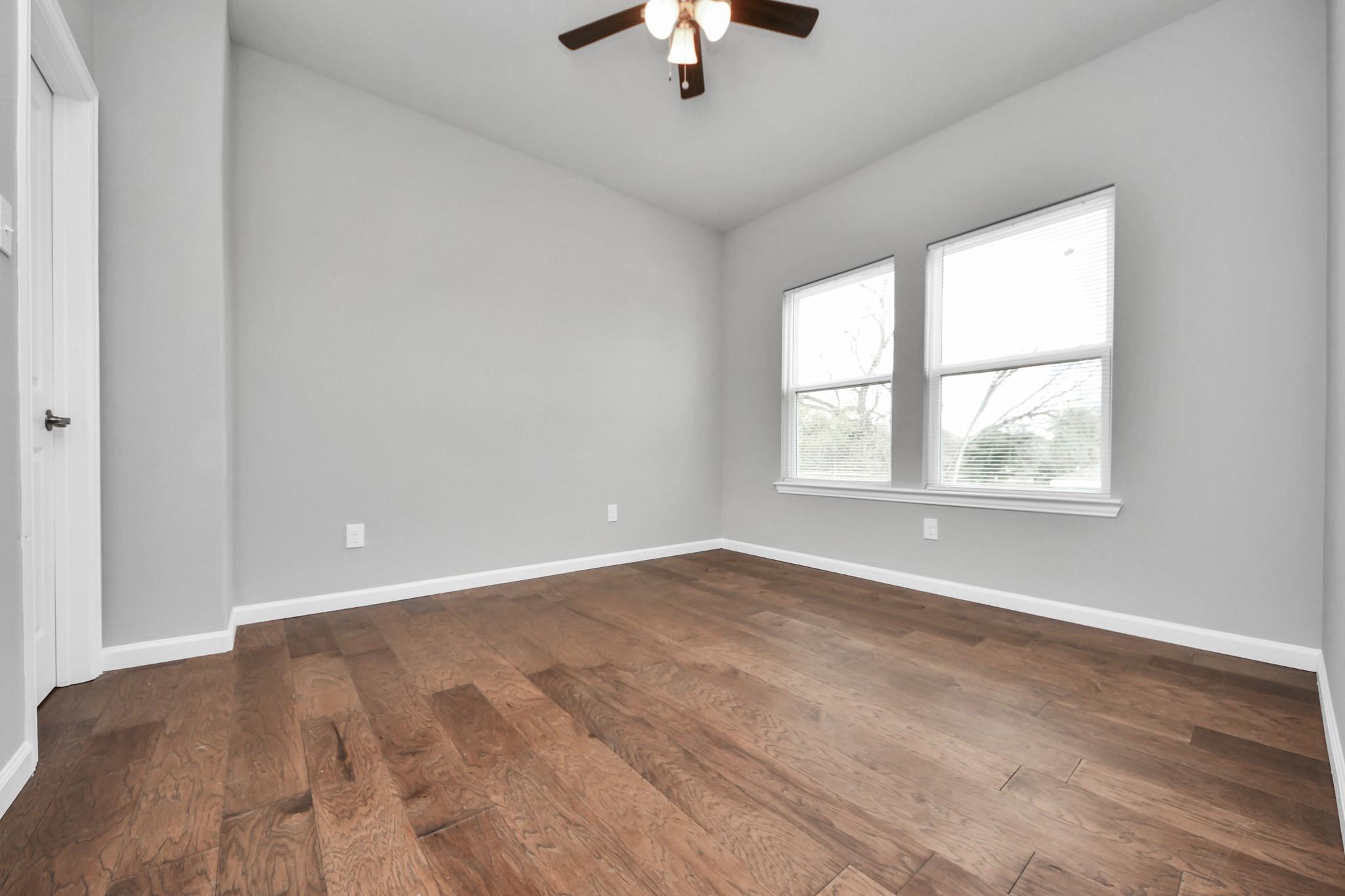 Empty bedroom with wood flooring, two windows with blinds, and a ceiling fan.