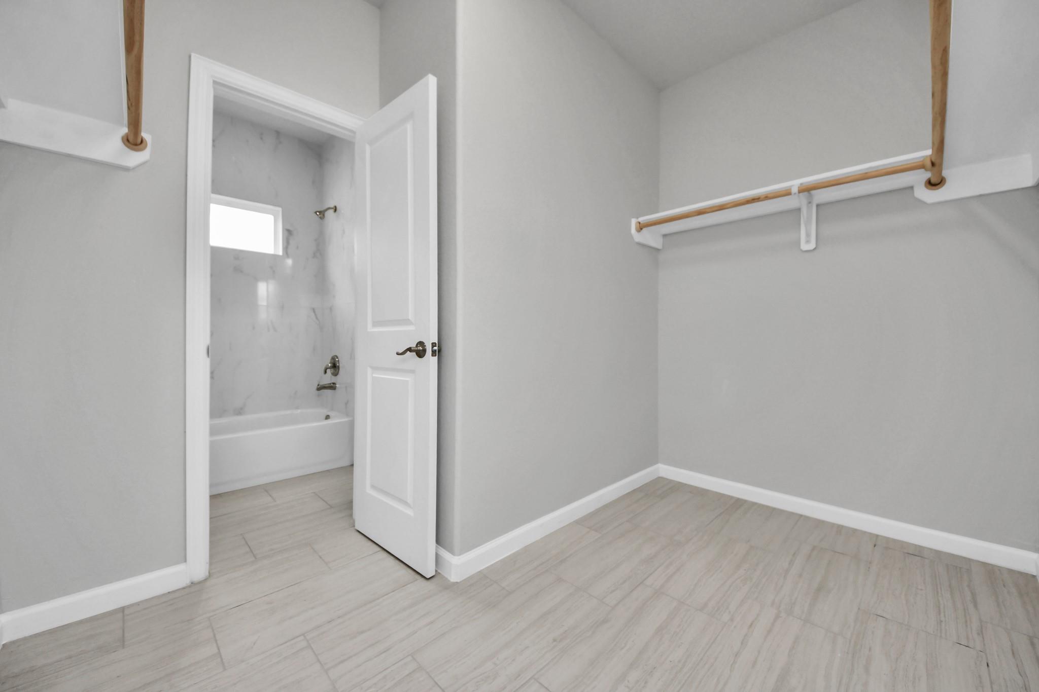 Empty walk-in closet with wooden hanging rods and an open doorway to a tiled bathroom.