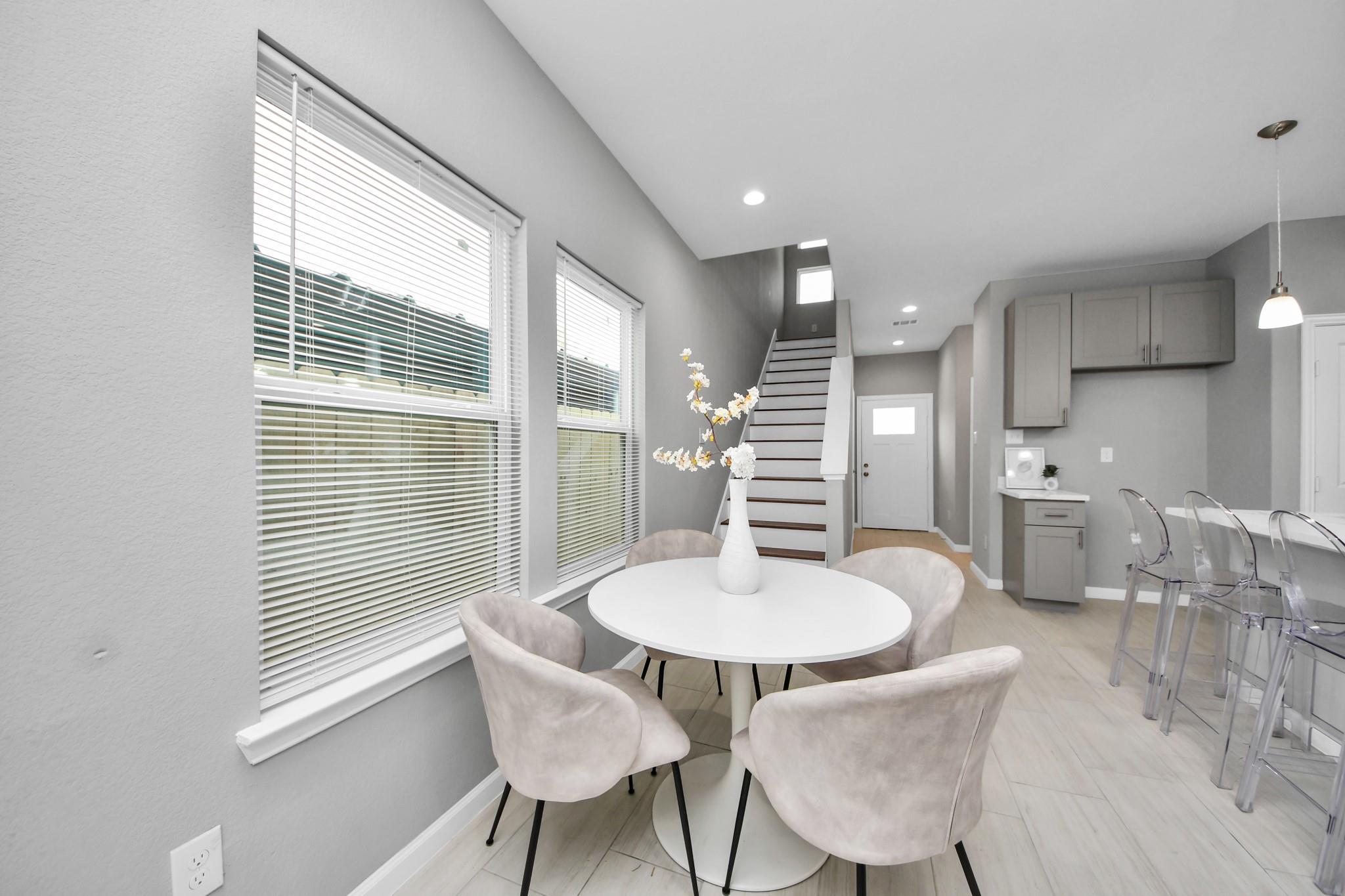 Dining area with a round white table beside tall windows, an open kitchen, and staircase.
