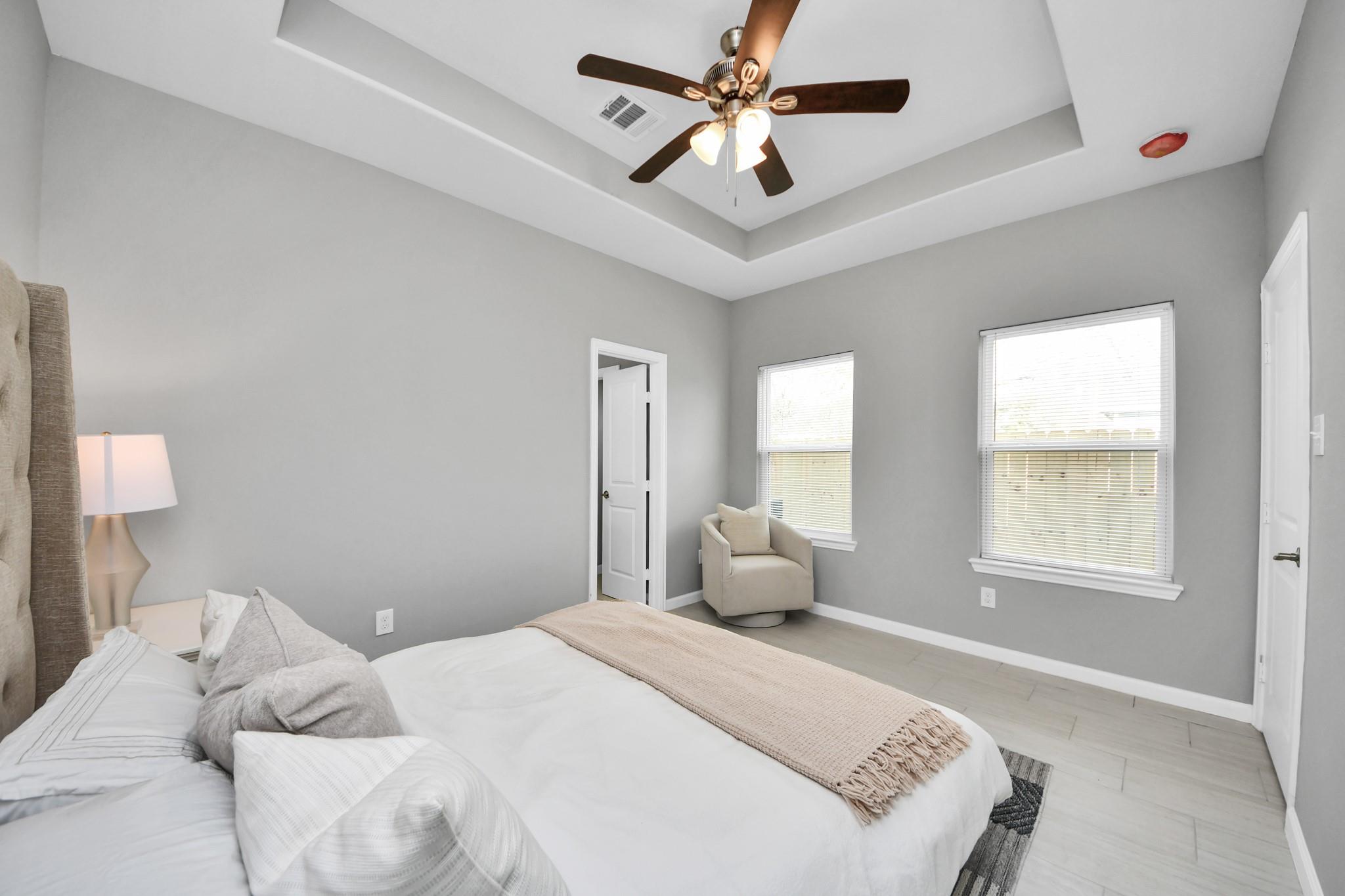 Staged bedroom with tray ceiling, ceiling fan, two windows, and upholstered armchair.