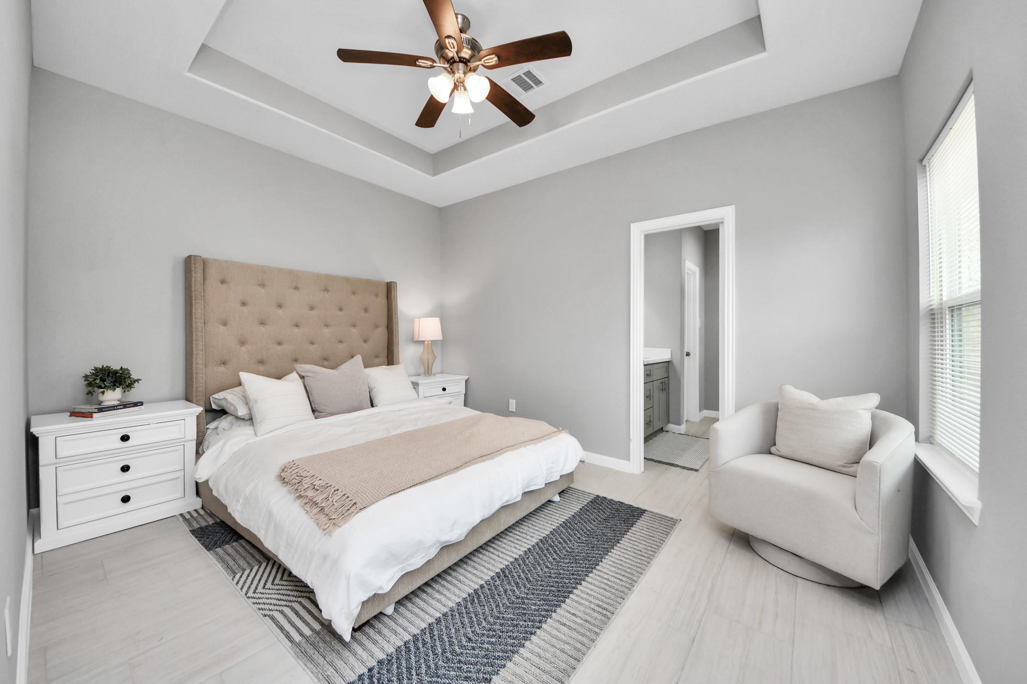 Bedroom with a tufted beige headboard, tray ceiling, ceiling fan, and armchair beside a bright window.
