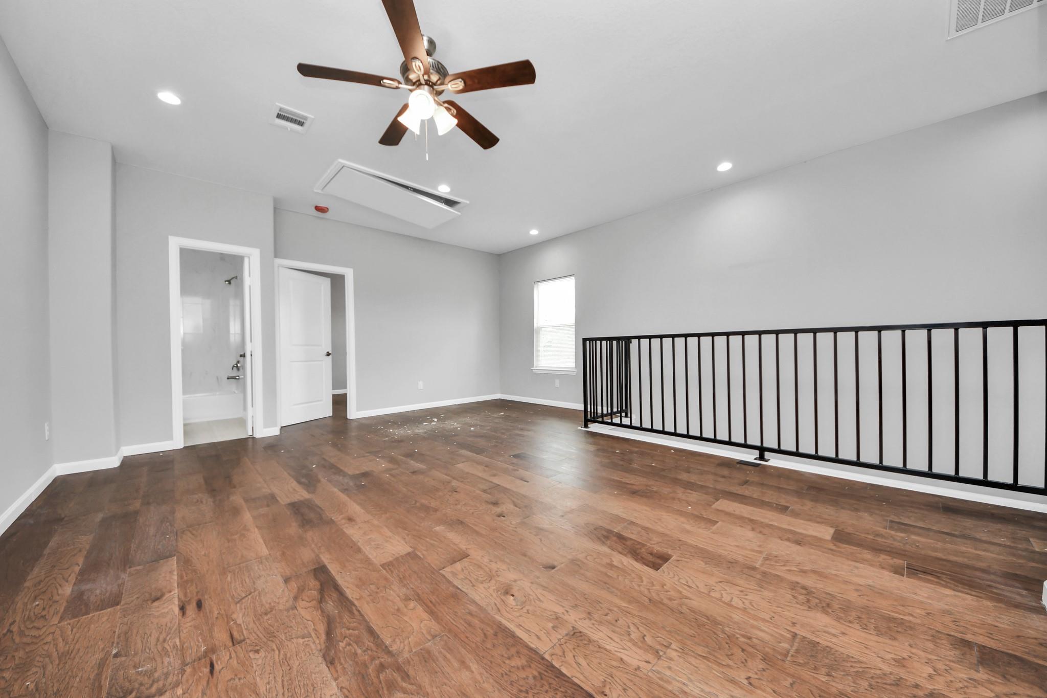 Upstairs loft with wood flooring, ceiling fan, and black railing beside a bathroom doorway.