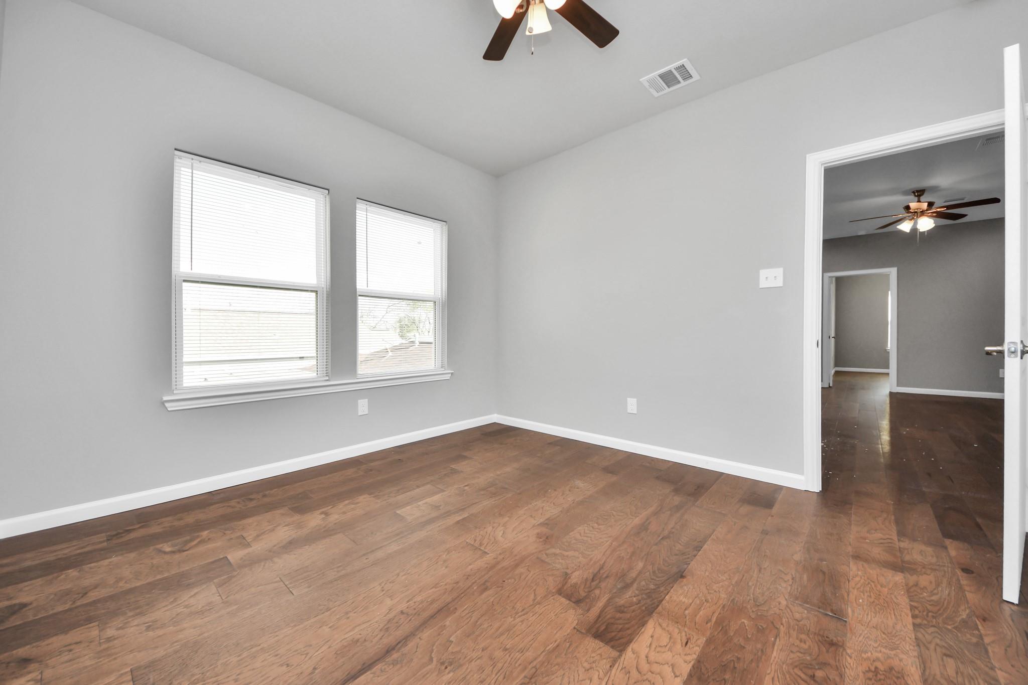 Empty room with hardwood floors, two windows, and a ceiling fan; doorway opens to another room.