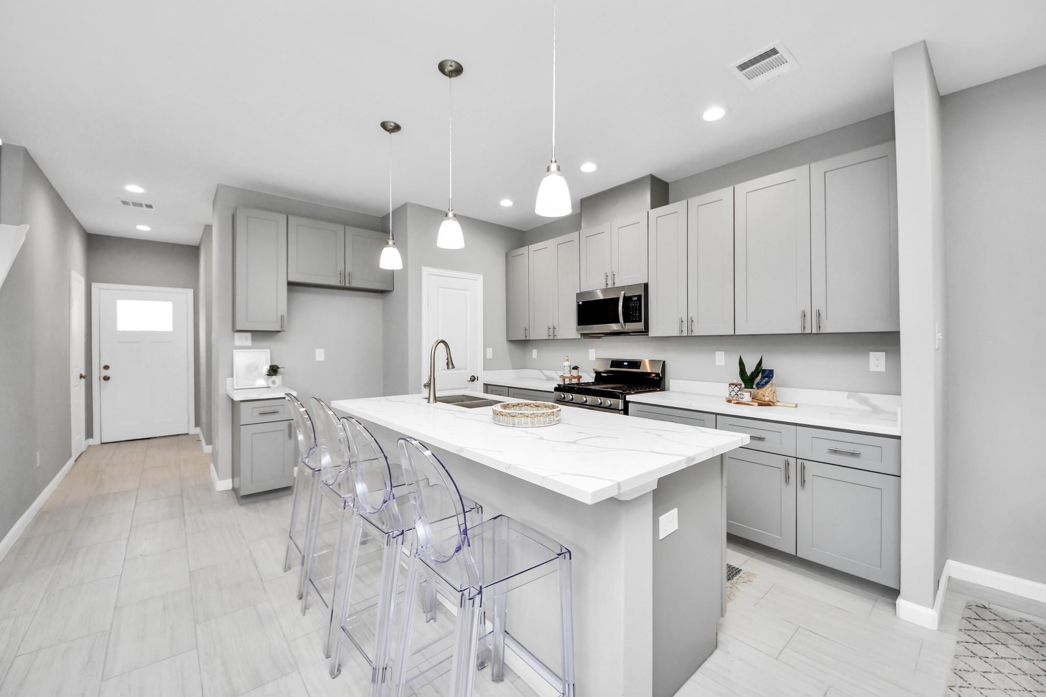 Modern kitchen with a large white island, clear bar stools, gray cabinets, and stainless steel appliances.