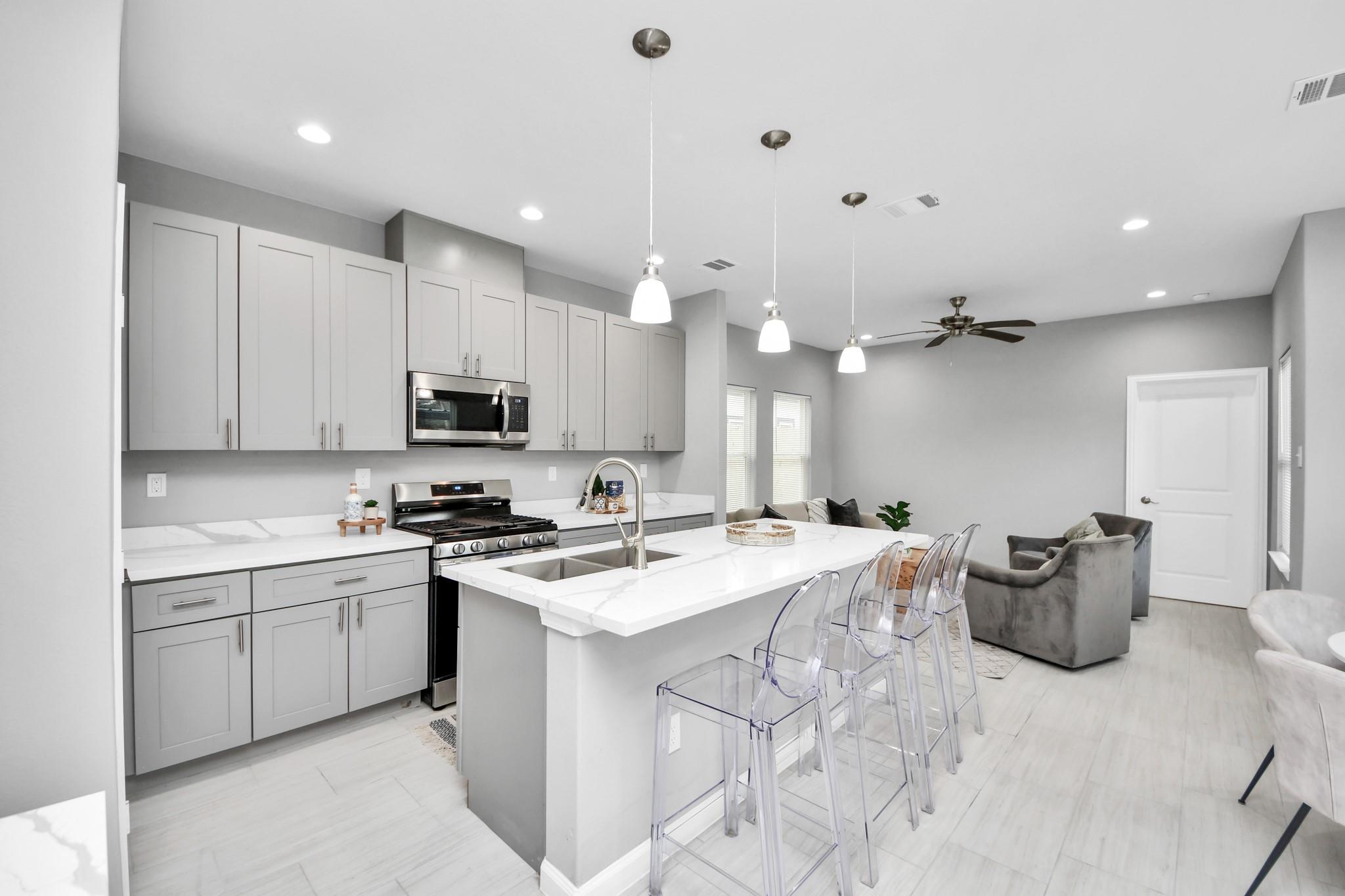 Open-plan kitchen with gray cabinets, stainless appliances, and a large island with clear bar stools.