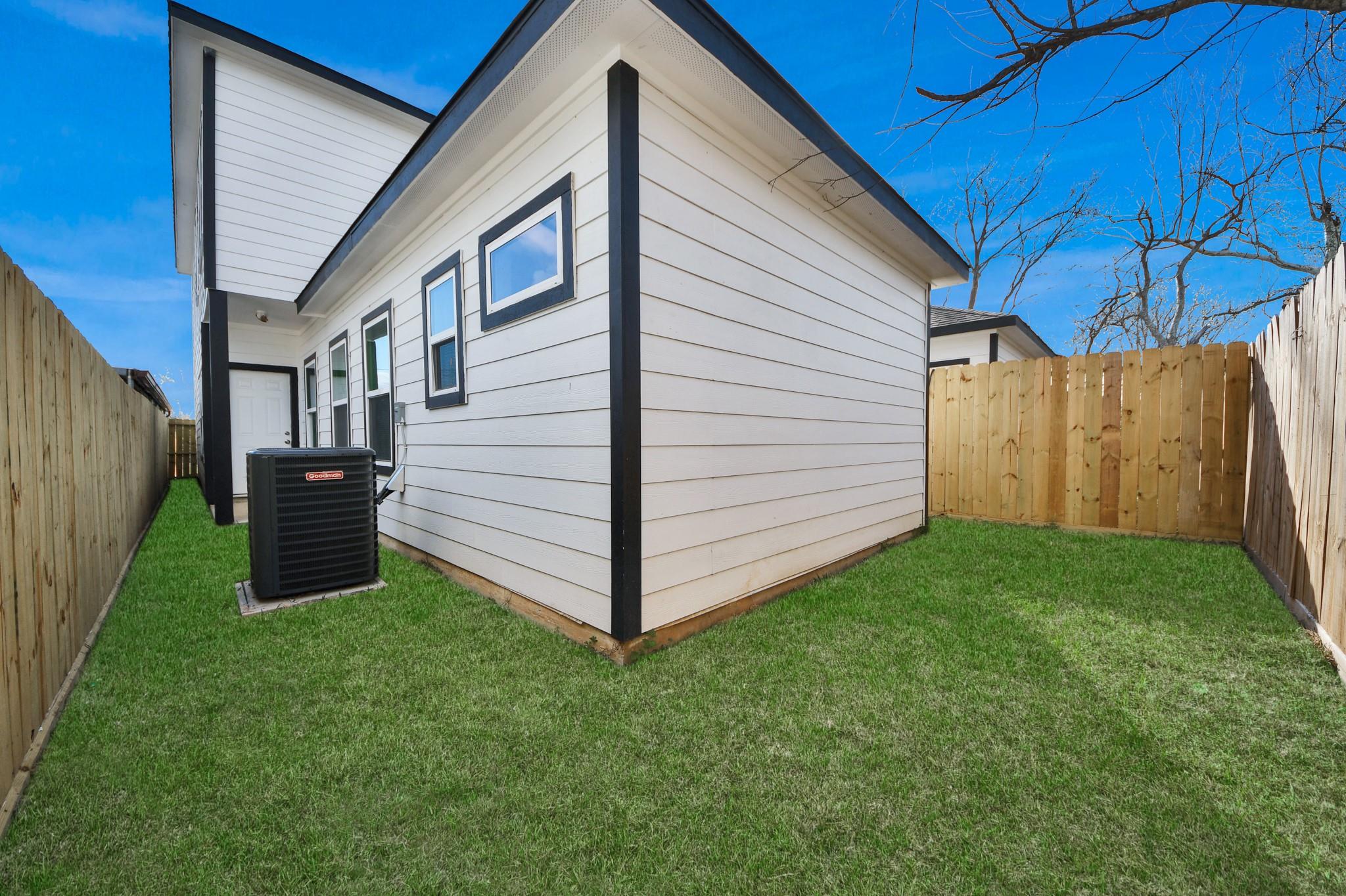 White house with black trim beside a fenced grassy yard and outdoor air-conditioning unit.