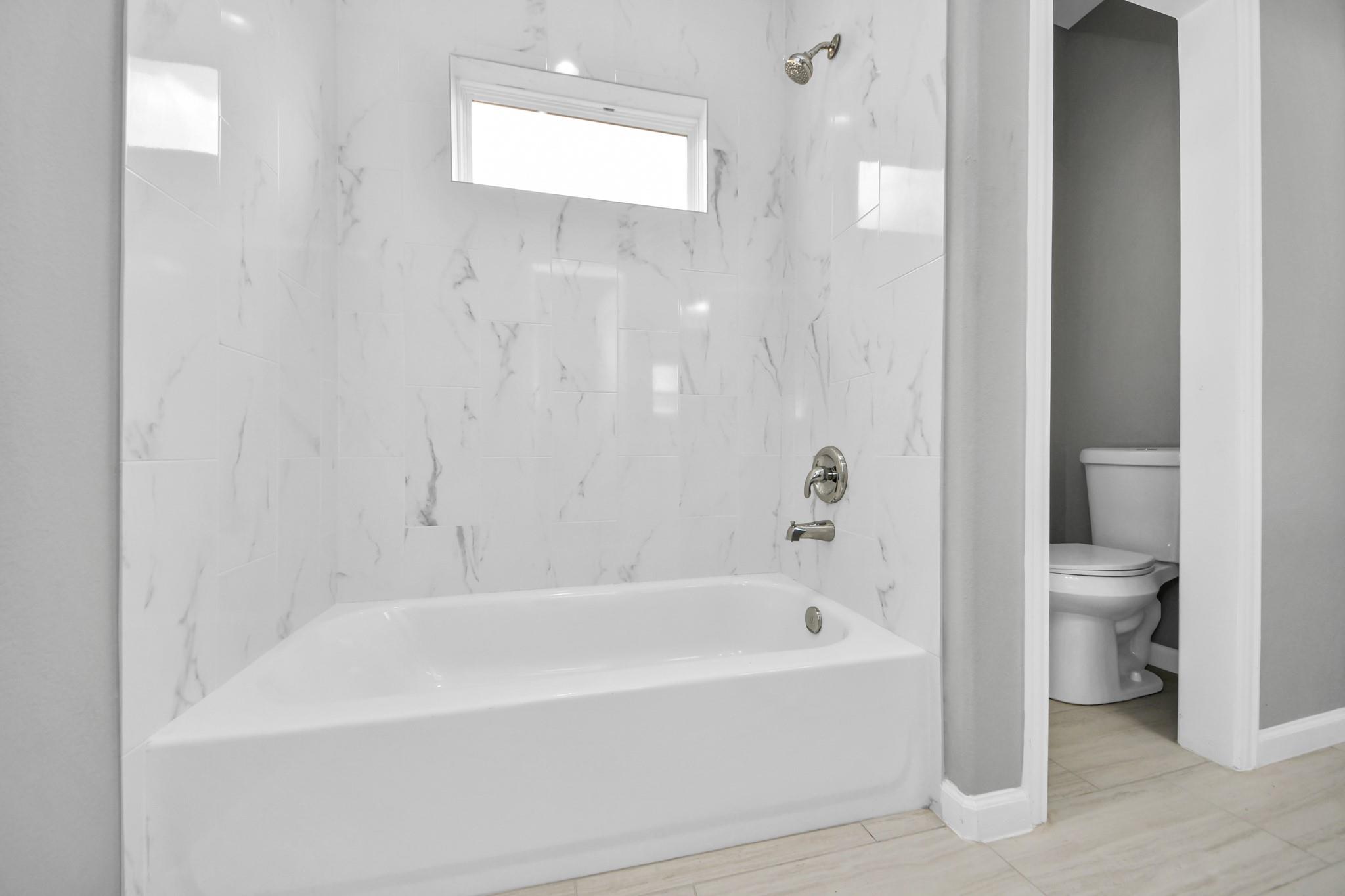 Bathroom with a white tub, marble-look tiled walls, and a toilet alcove.
