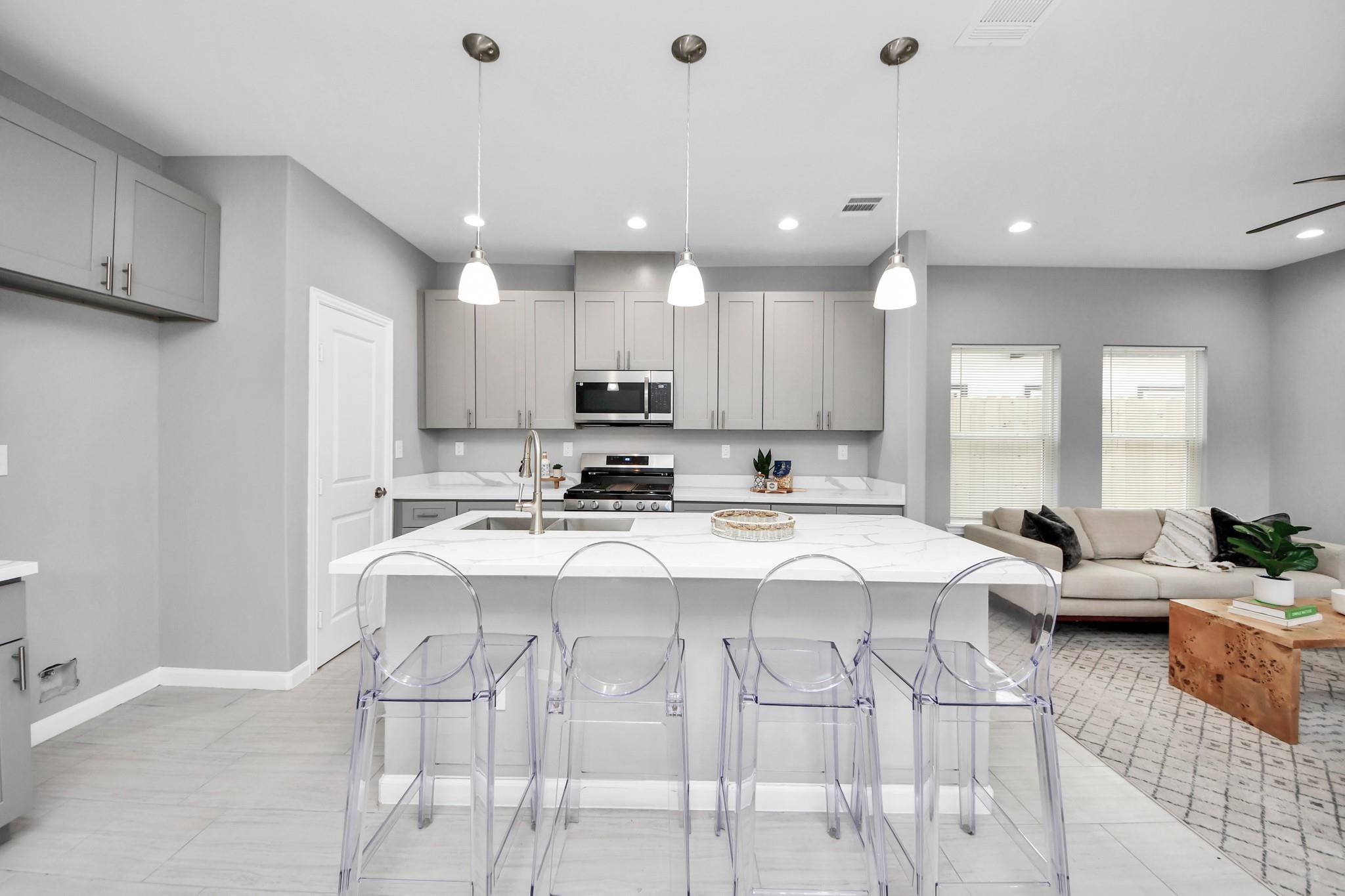 Open kitchen with a large white island and clear stools adjoining a living area with a beige sofa.
