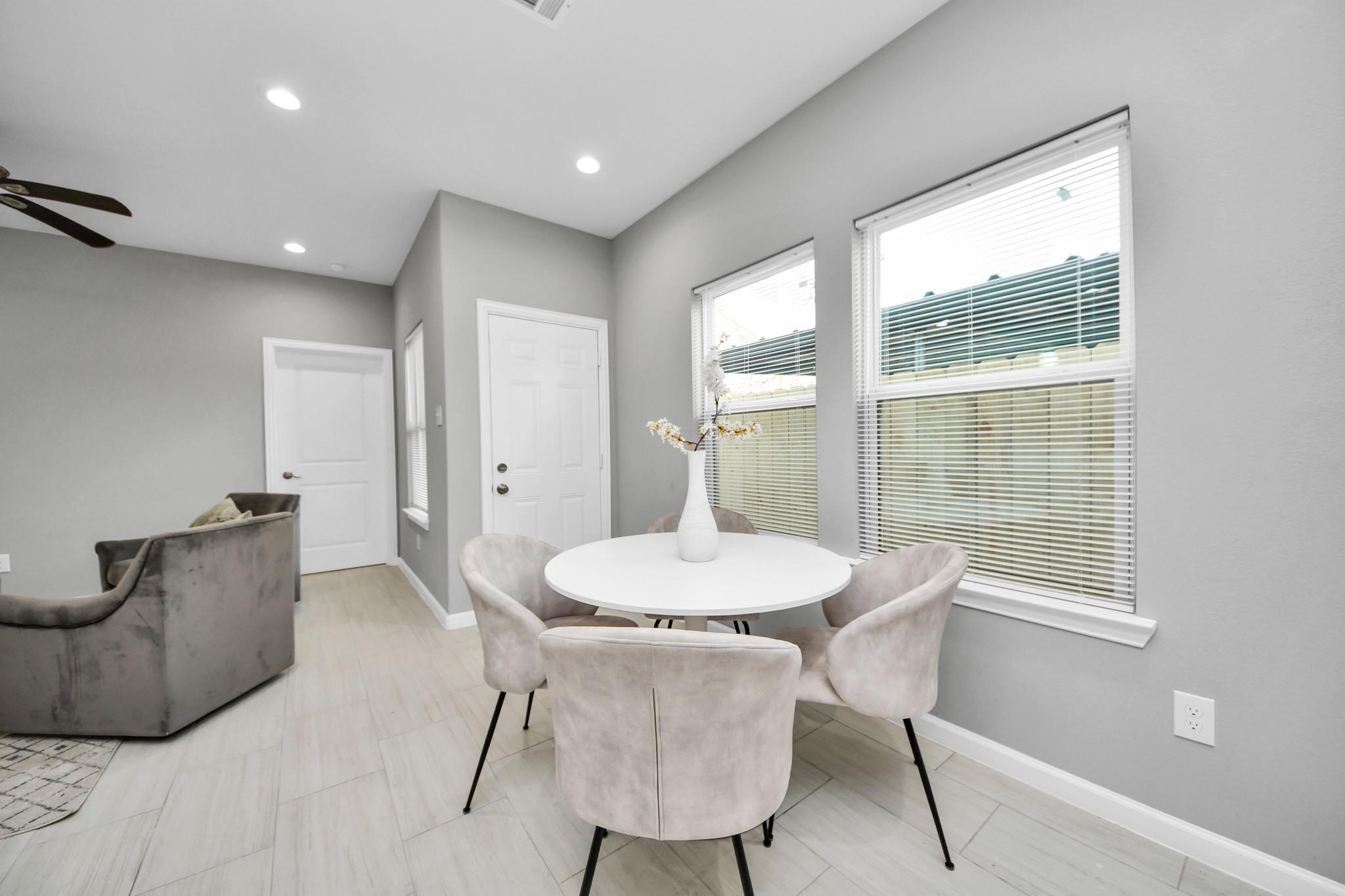 Dining nook with a round white table and upholstered chairs beside large windows with blinds.