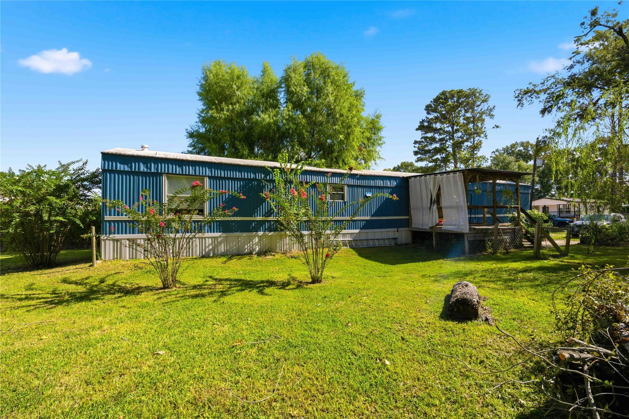 Blue manufactured home with a curtained covered deck, flowering shrubs, and a broad grassy yard.