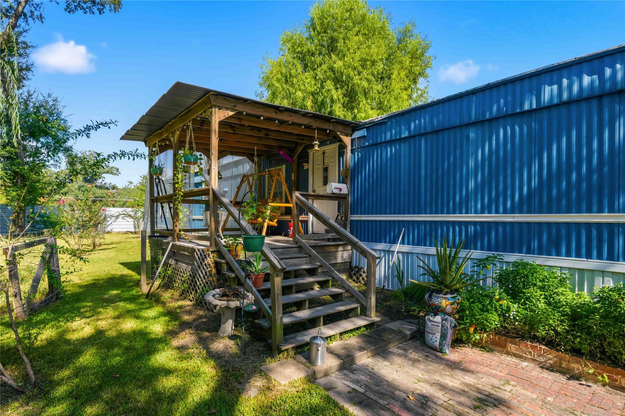 Blue-sided home with a covered wooden porch, broad steps, hanging plants, and a brick patio.