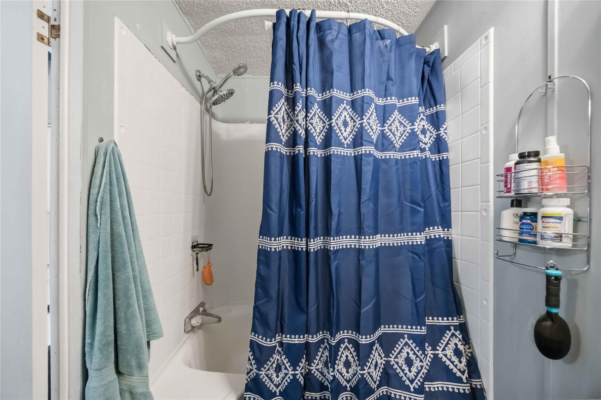 Bathroom shower and tub with white tile, a blue patterned curtain, and toiletries on a wall rack.