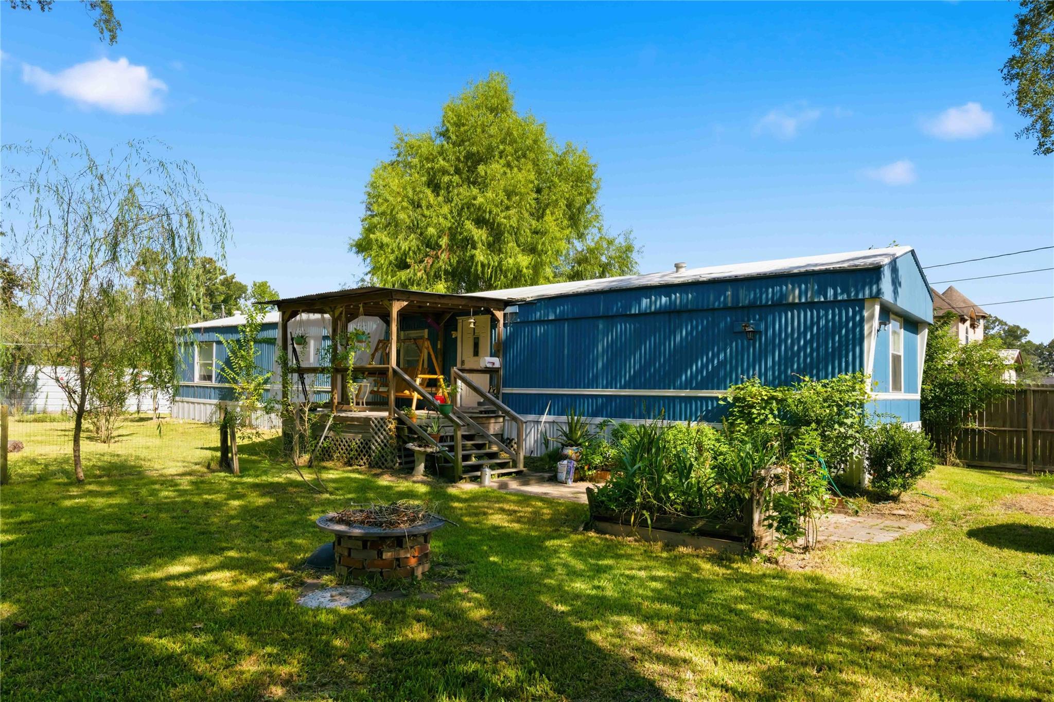 Blue manufactured home with a covered porch, garden beds, and a brick fire pit in the yard.