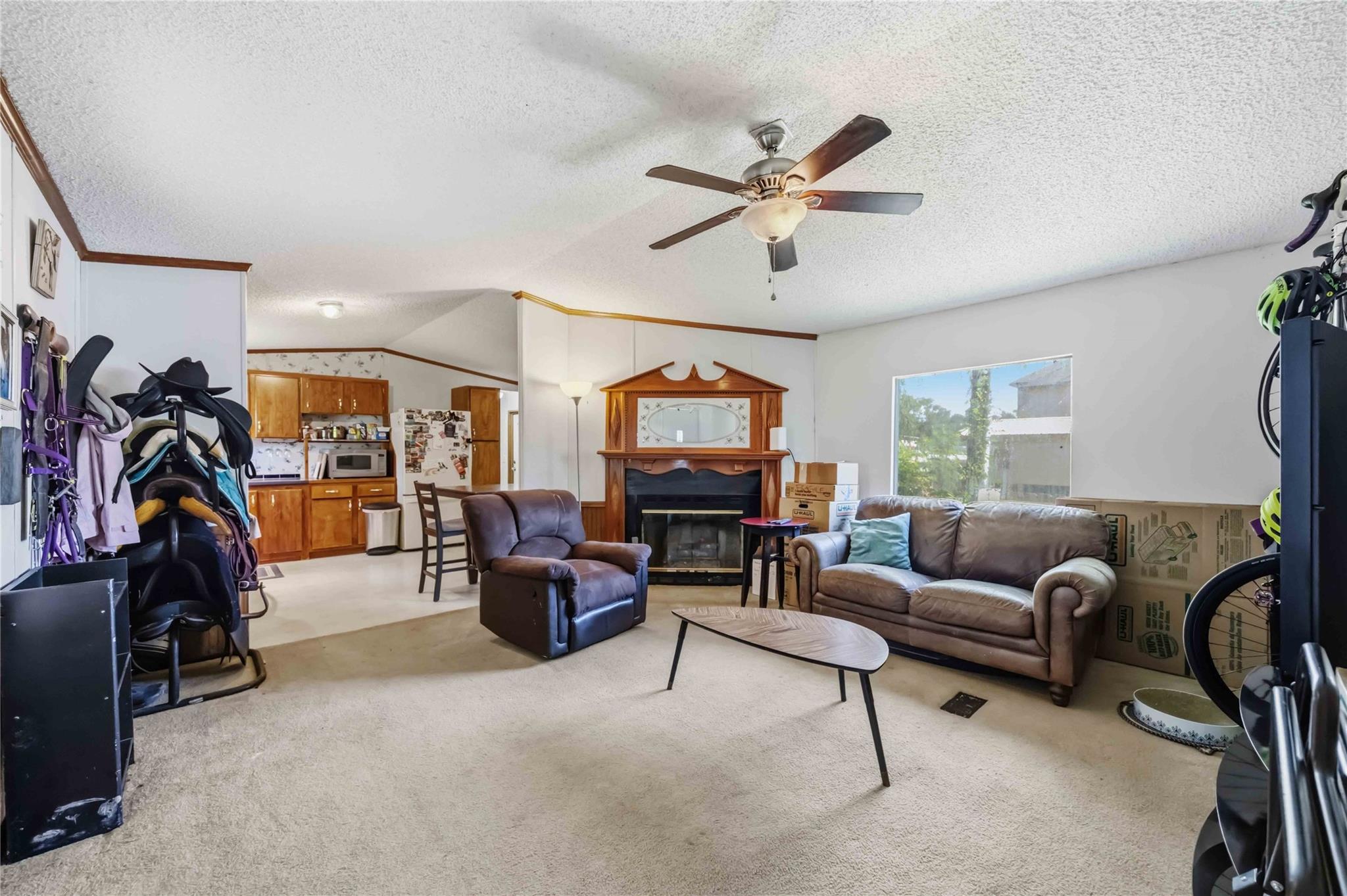 Living room with a wood-trimmed fireplace, carpet, and open kitchen; boxes and bicycles line the walls.