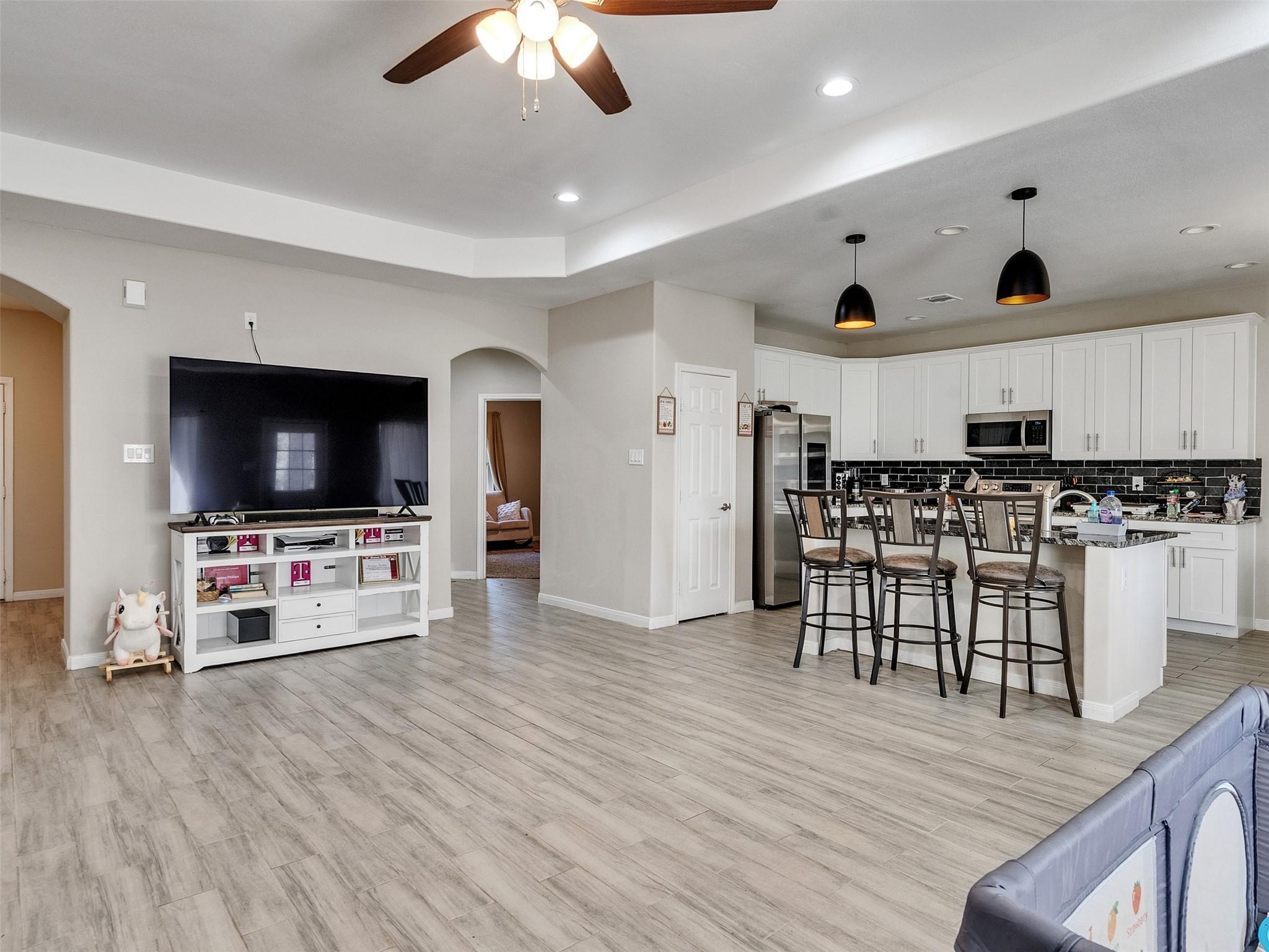 Open-plan living room and kitchen with a large island, white cabinets, black backsplash, and gray tile flooring.