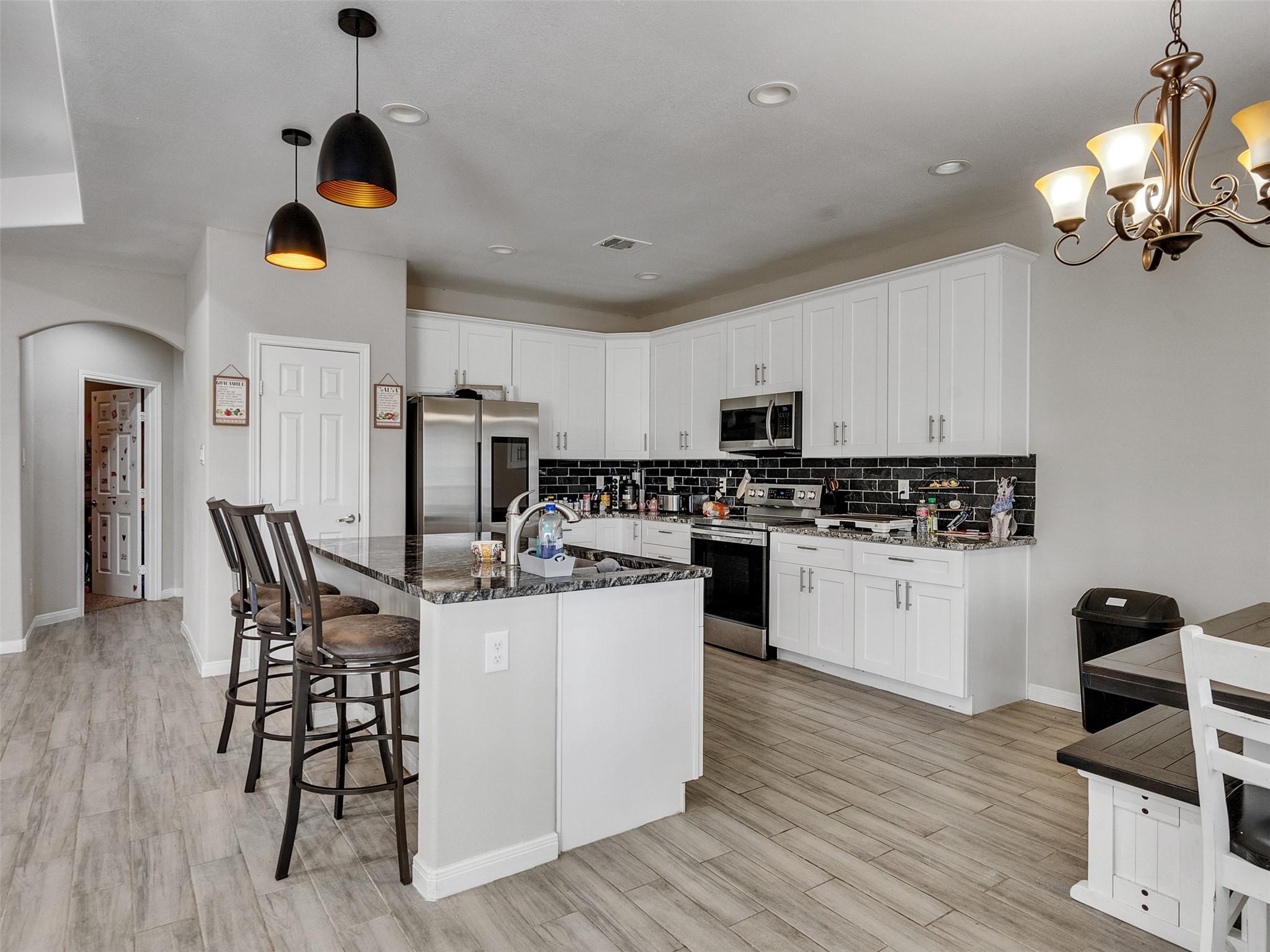 Kitchen with white cabinetry, black tile backsplash, stainless appliances, and a granite island with bar stools.