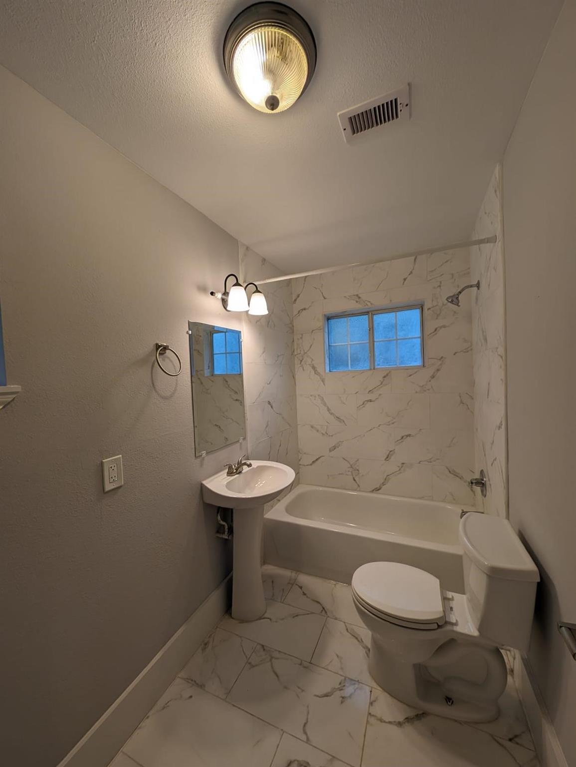 Bathroom with a tub and shower, pedestal sink, toilet, and marble-look tile.