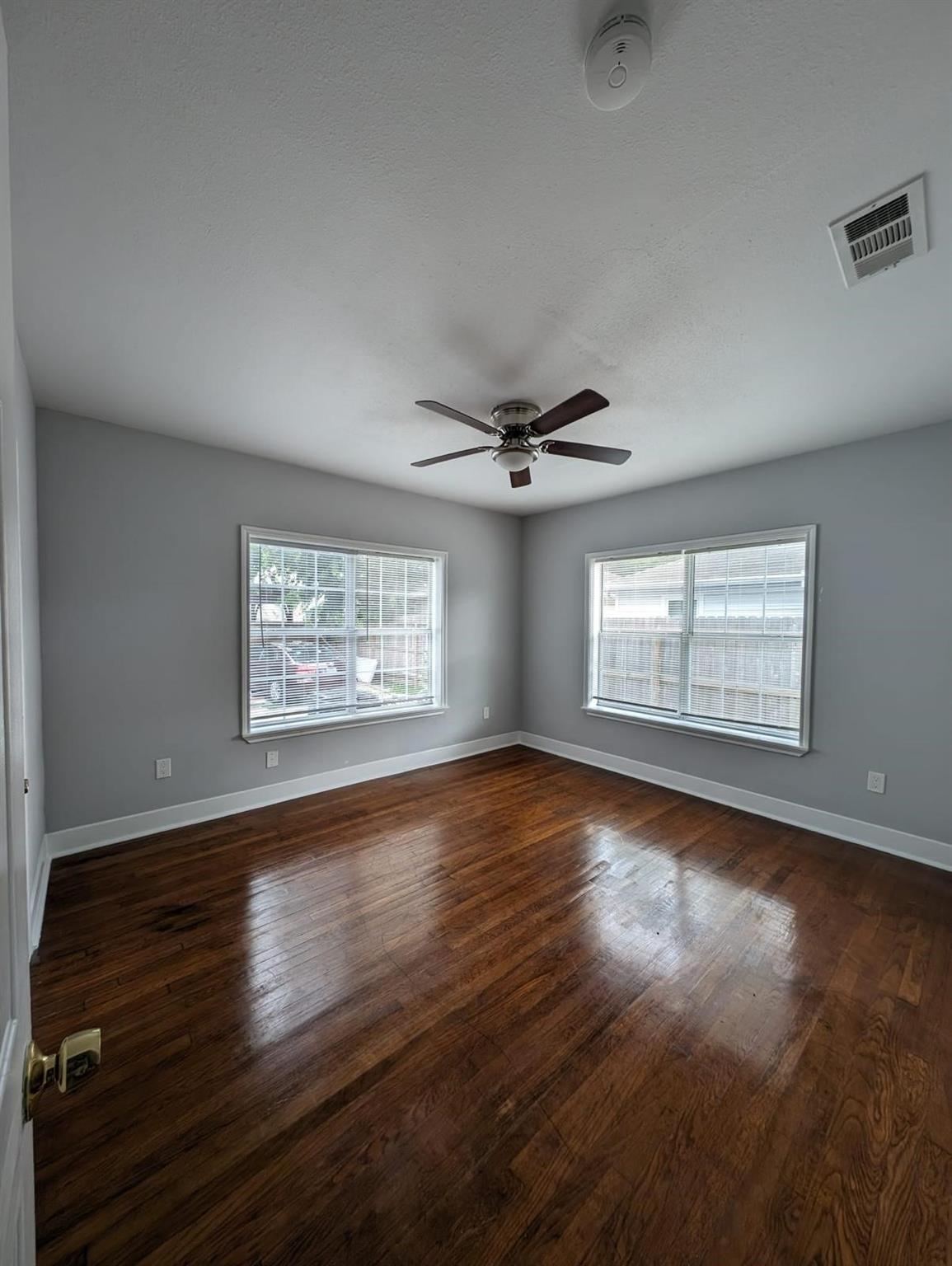 Empty bedroom with dark hardwood floors, gray walls, two windows, and a ceiling fan.