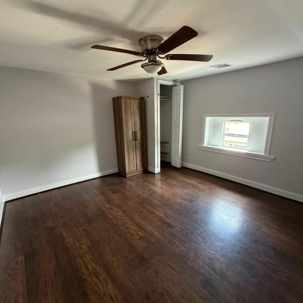 Empty bedroom with dark wood floors, ceiling fan, small window, and wooden wardrobe.