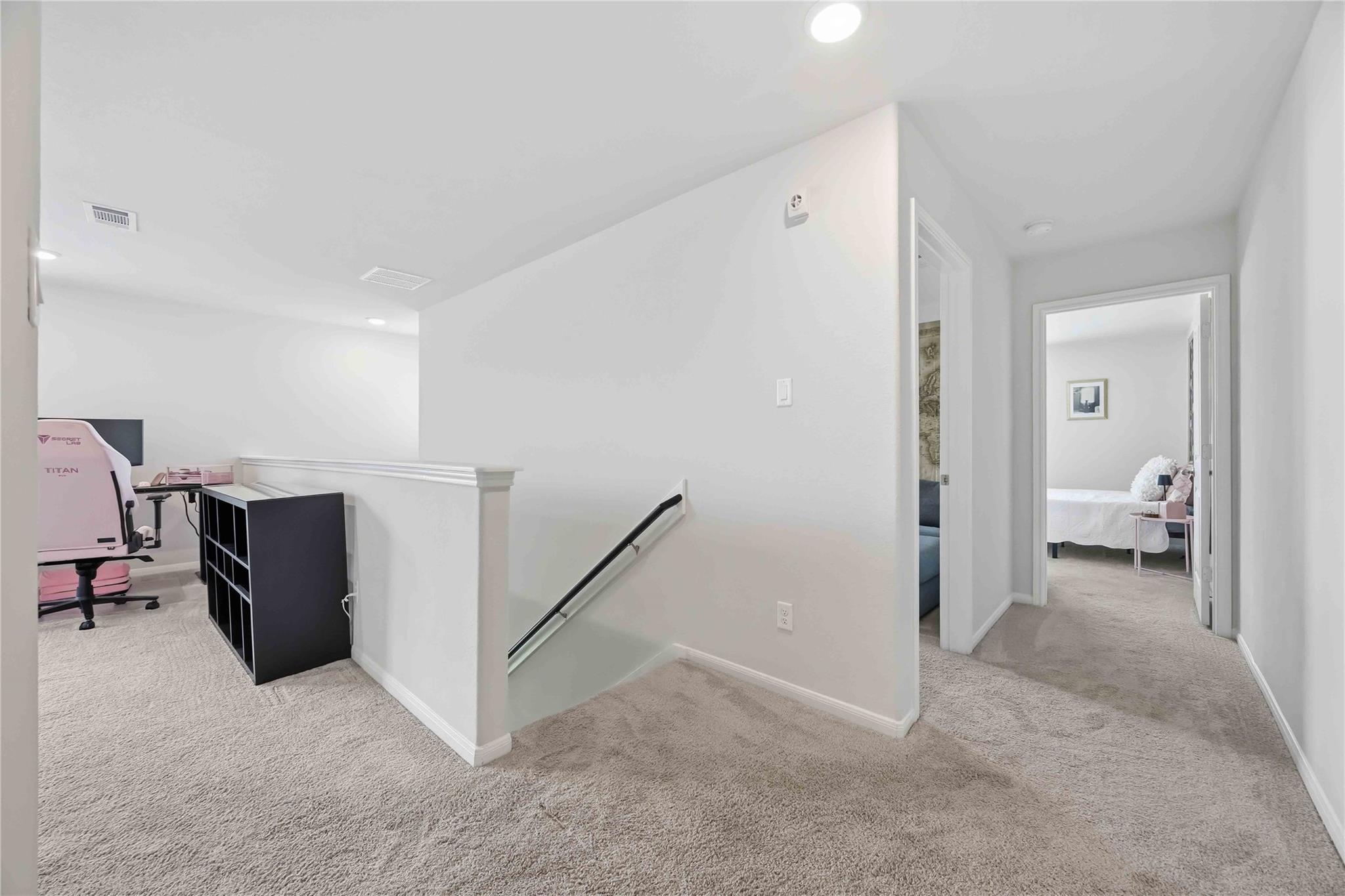 Carpeted upstairs landing with a stairwell, pink gaming chair, and bedroom visible through the hallway.