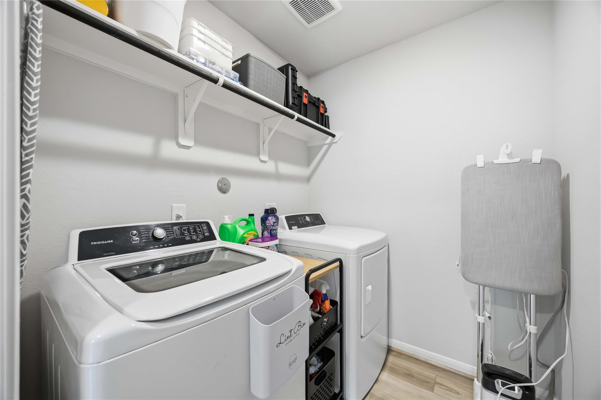 Laundry room with a top-loading washer and dryer, wall storage shelf, and folding ironing board.