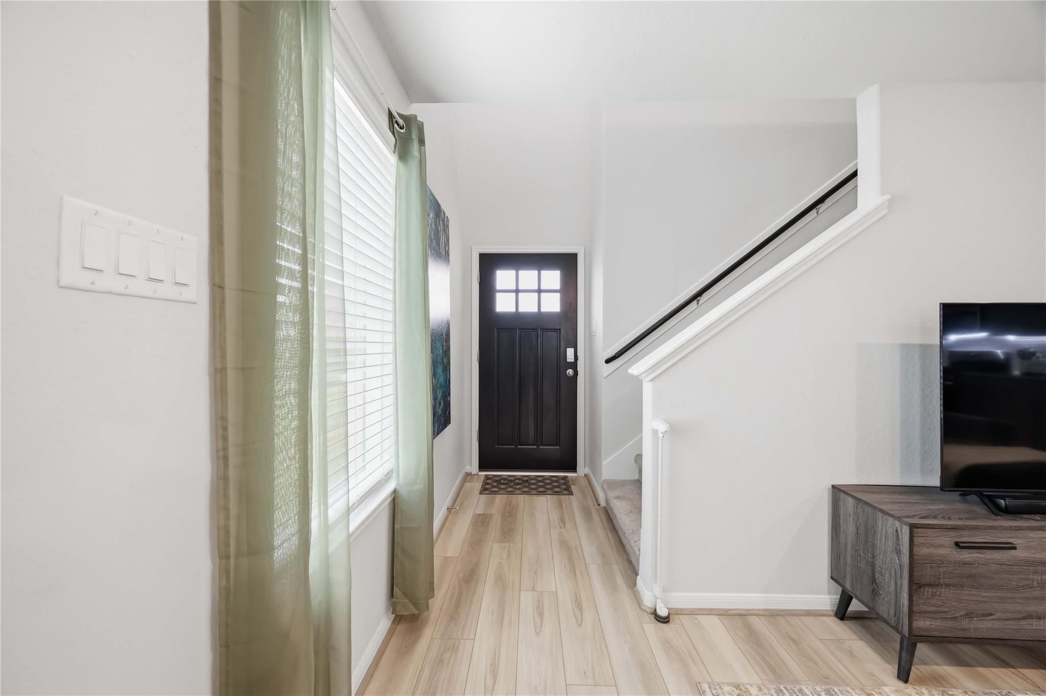 Entryway with a dark front door, pale wood floors, and staircase beside a television console.