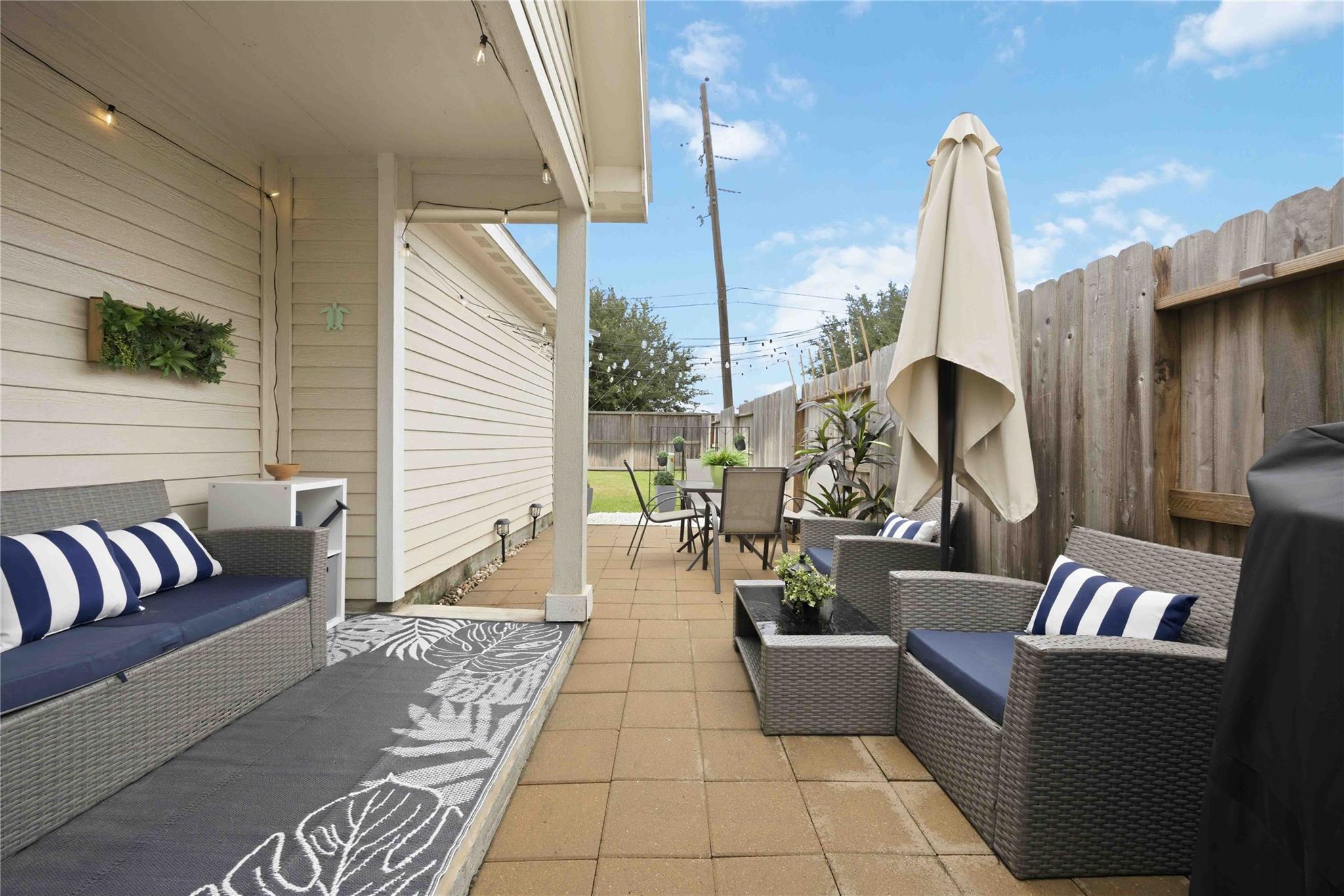 Covered backyard patio with wicker seating, striped cushions, and a closed umbrella.