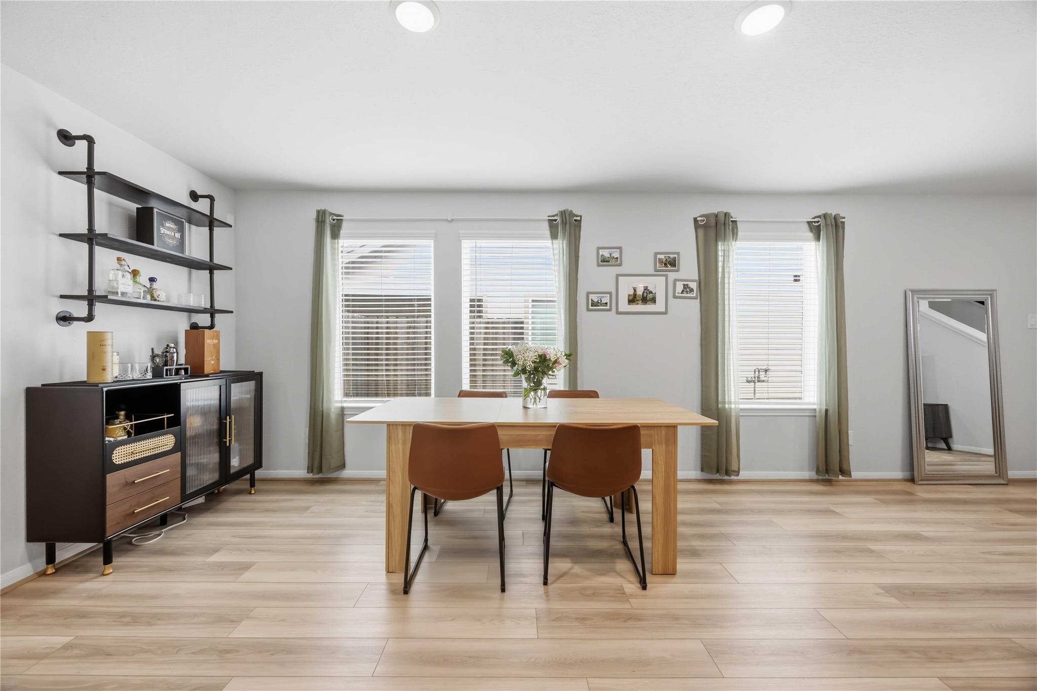 Dining room with a light wood table, brown chairs, three curtained windows, and dark shelving.
