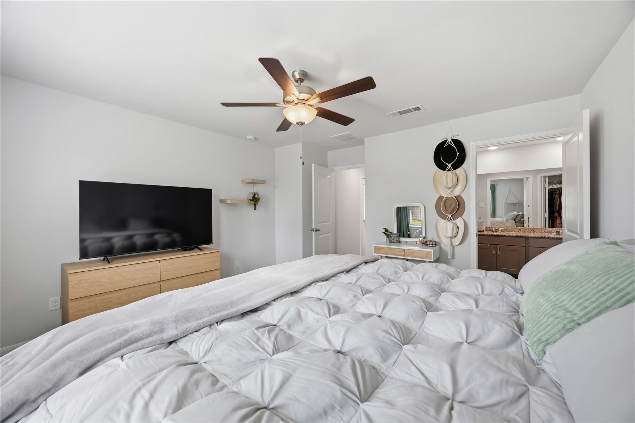 Furnished bedroom with a tufted white bed, ceiling fan, and open doorway to an en-suite bathroom.