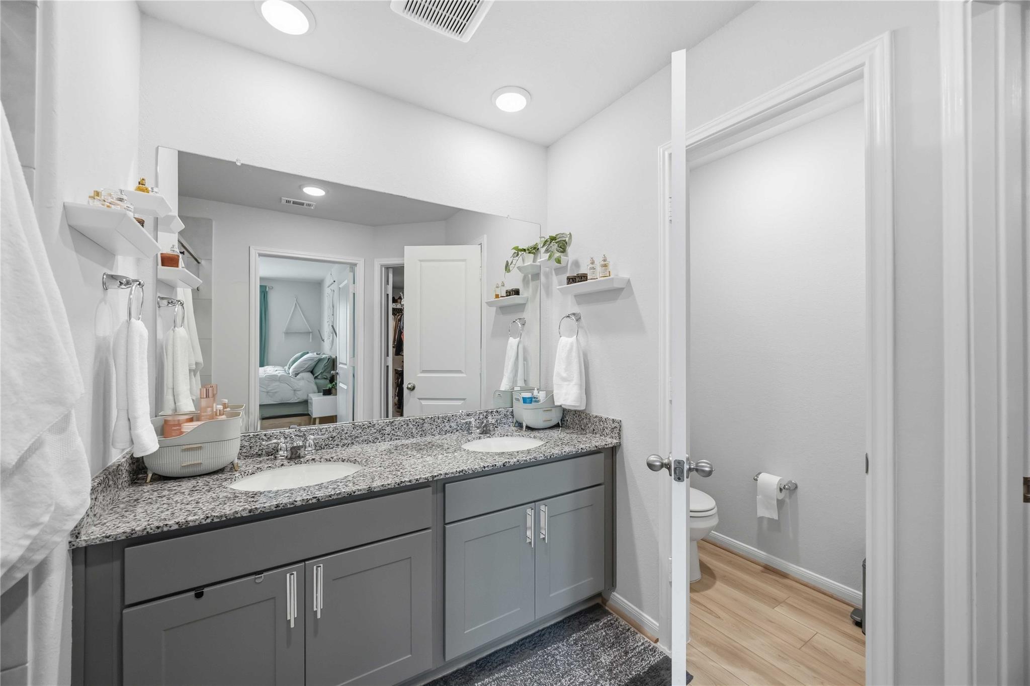 Bathroom with a gray double vanity and granite countertop, mirrored above two sinks; doorway opens to a separate toilet area.