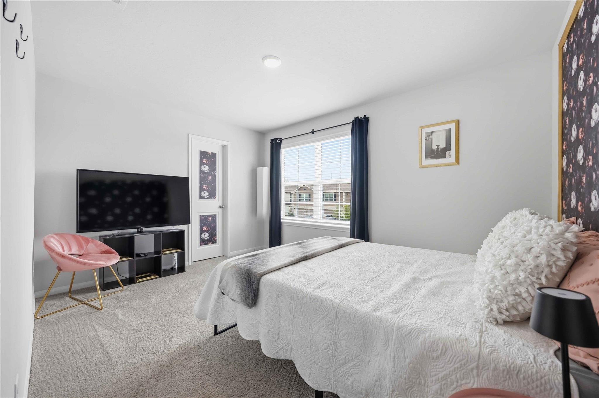 Staged bedroom with a white quilted bed, pink chair, television, and window framed by navy curtains.