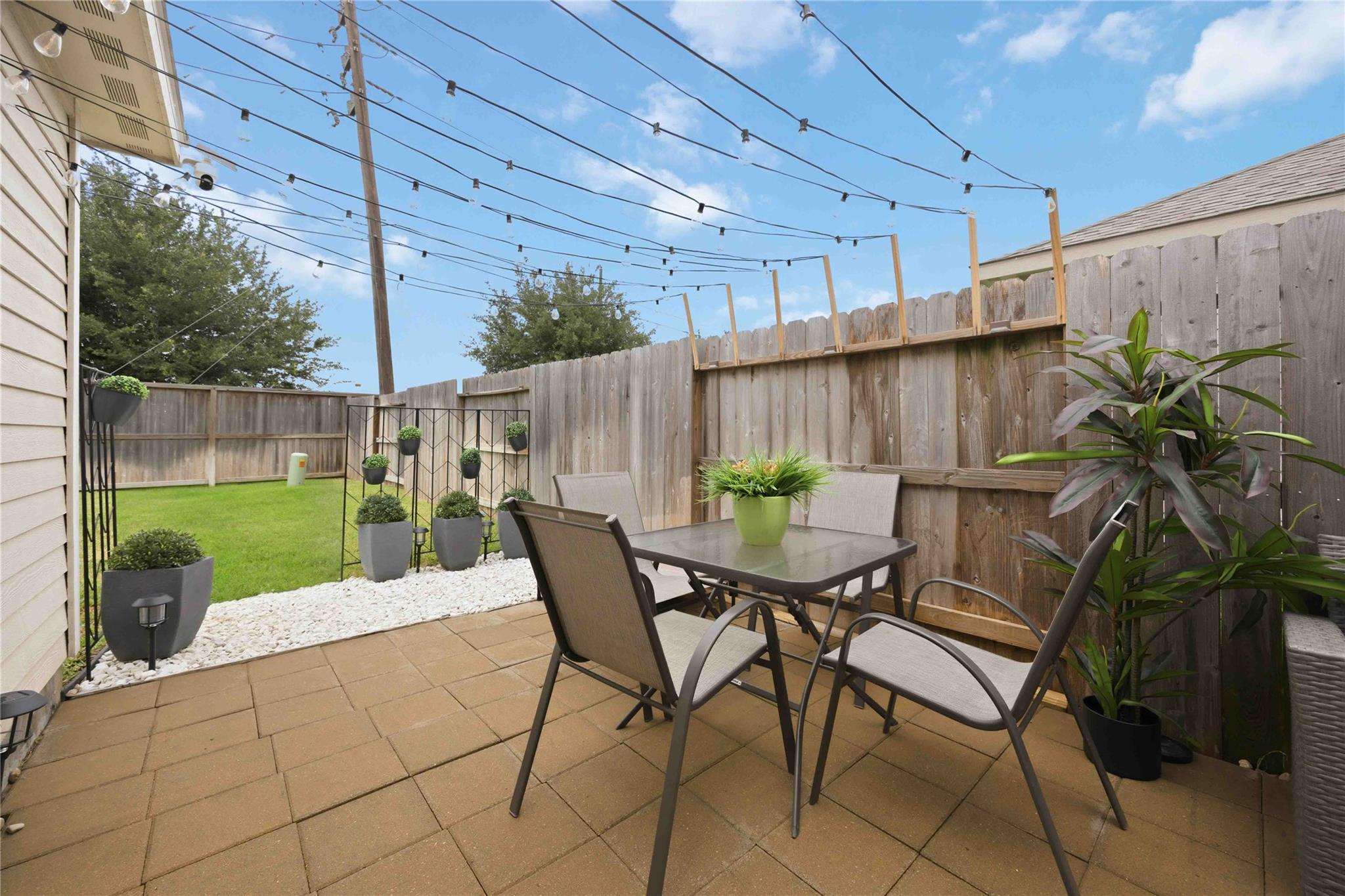 Backyard patio with a paver dining area, potted plants, string lights, and a fenced lawn.