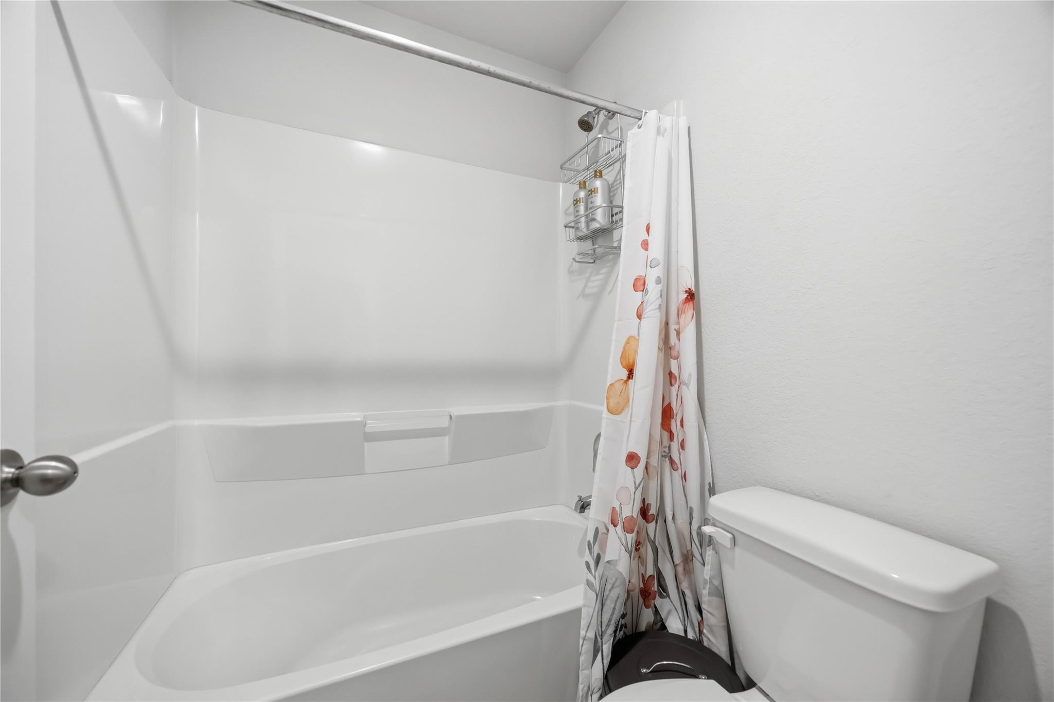 Bathroom with a white bathtub and shower surround, floral curtain, and toilet.