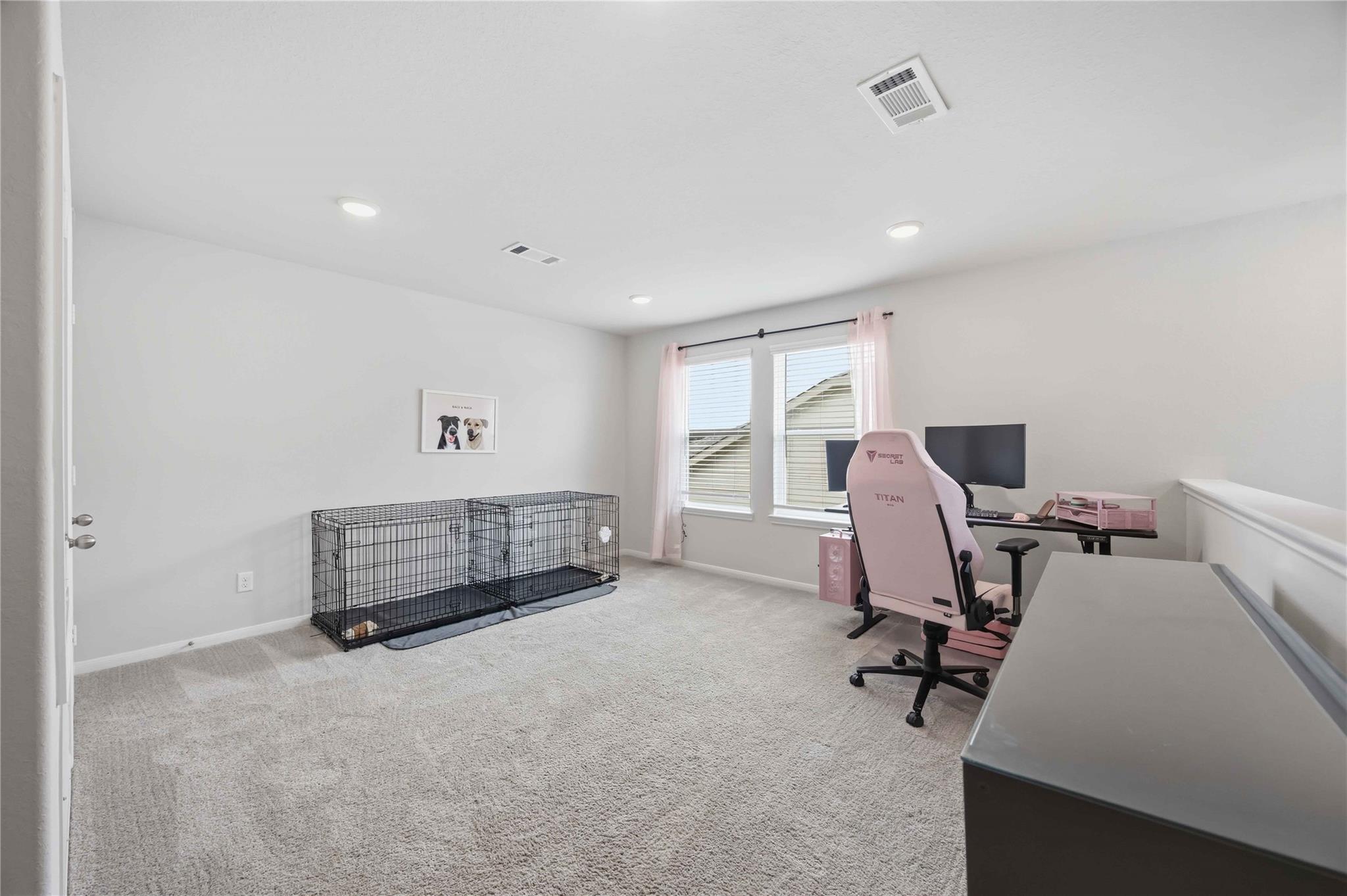 Carpeted home office with dog crates, dual monitors, and a pink gaming chair.