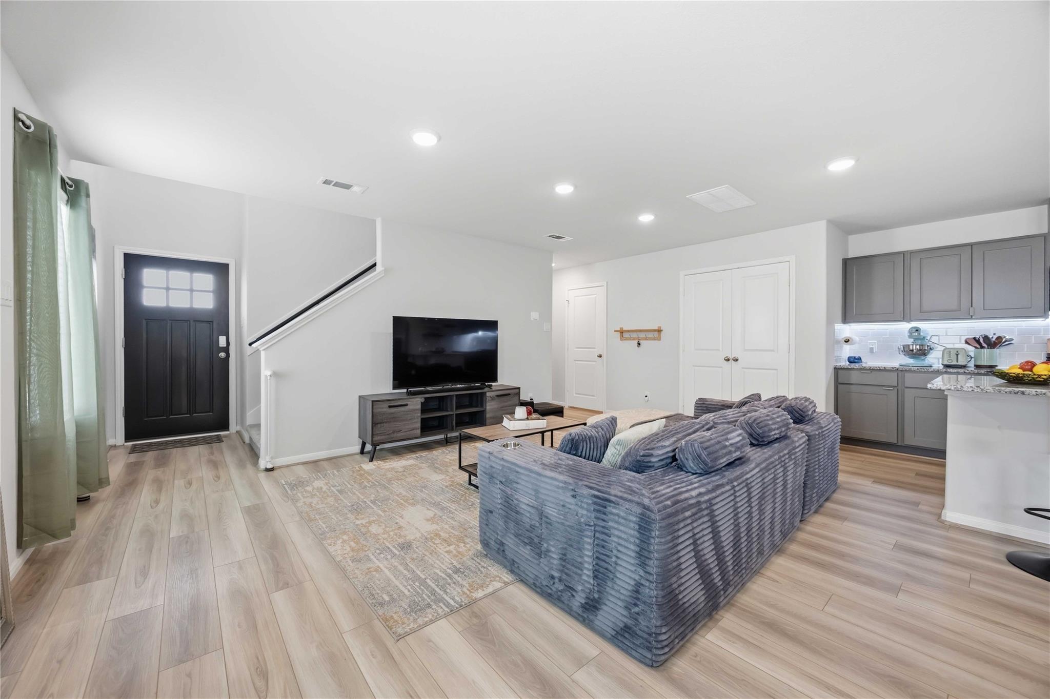 Open-plan living room with a gray sectional sofa, television, staircase, and adjoining kitchen.