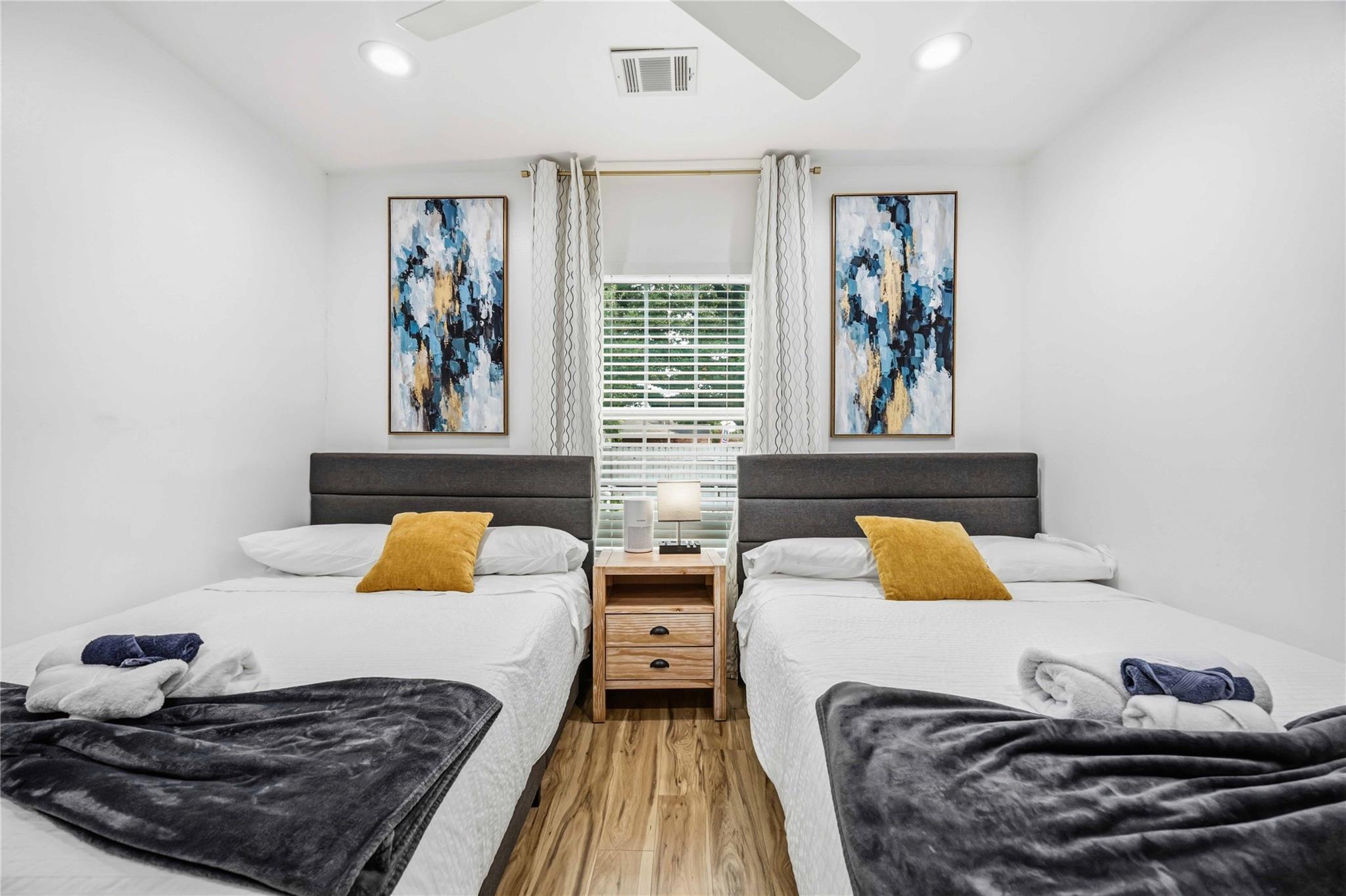 Bedroom with two double beds, mustard pillows, and abstract artwork flanking a curtained window.