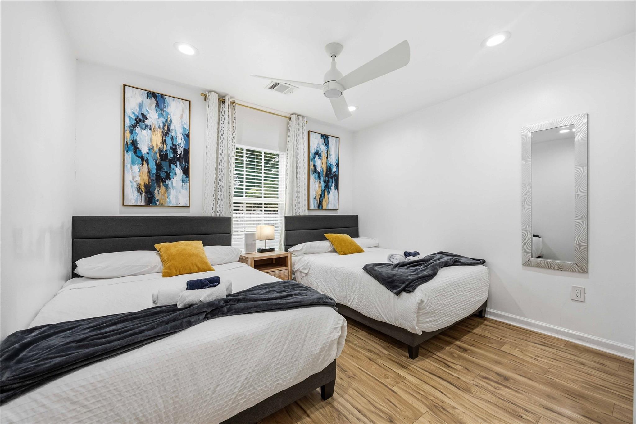 Bedroom with two beds, mustard-yellow pillows, abstract blue artwork, and wood-look flooring.