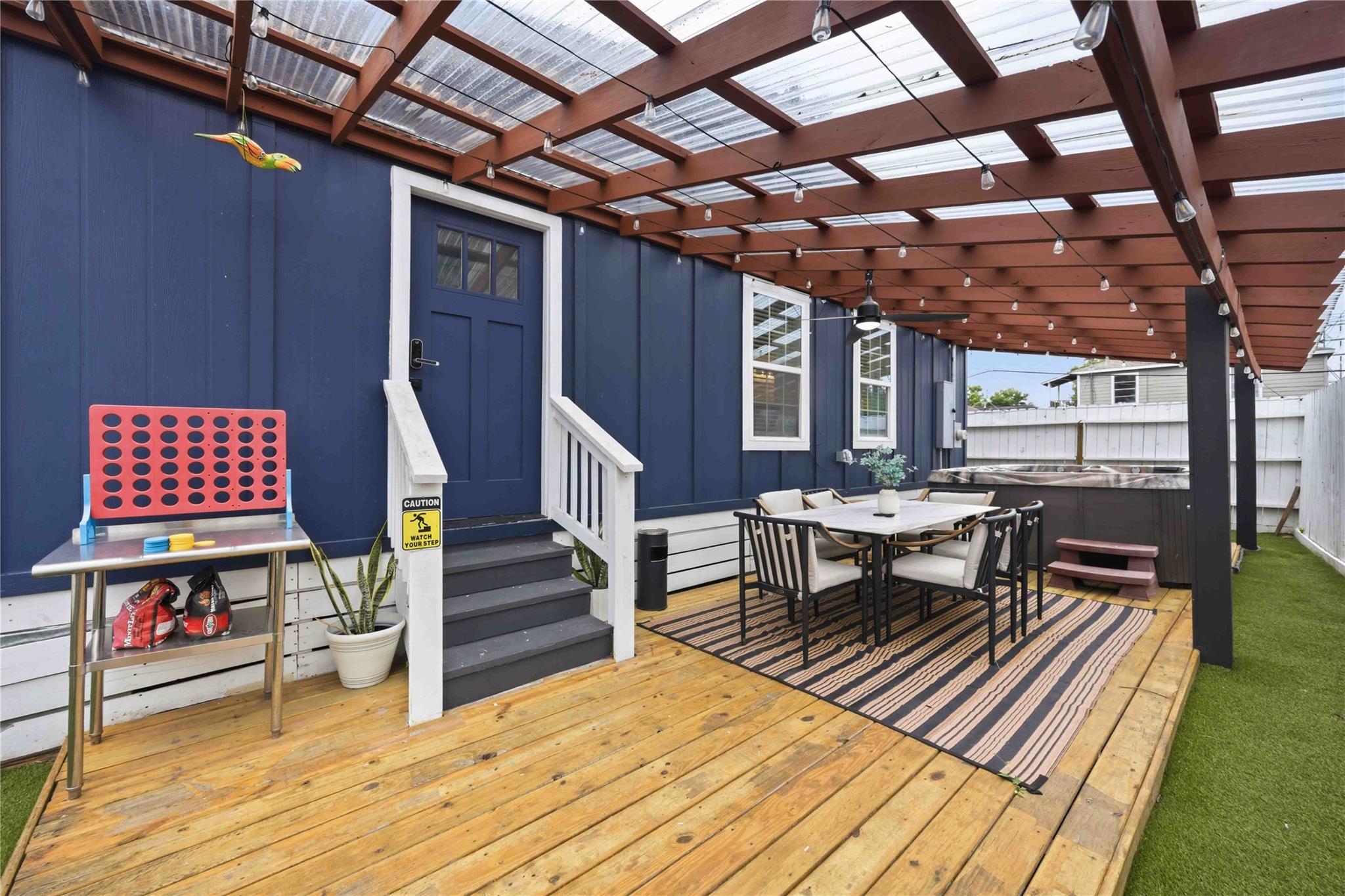 Covered deck with a wood pergola, outdoor dining set, and hot tub beside a dark blue house.