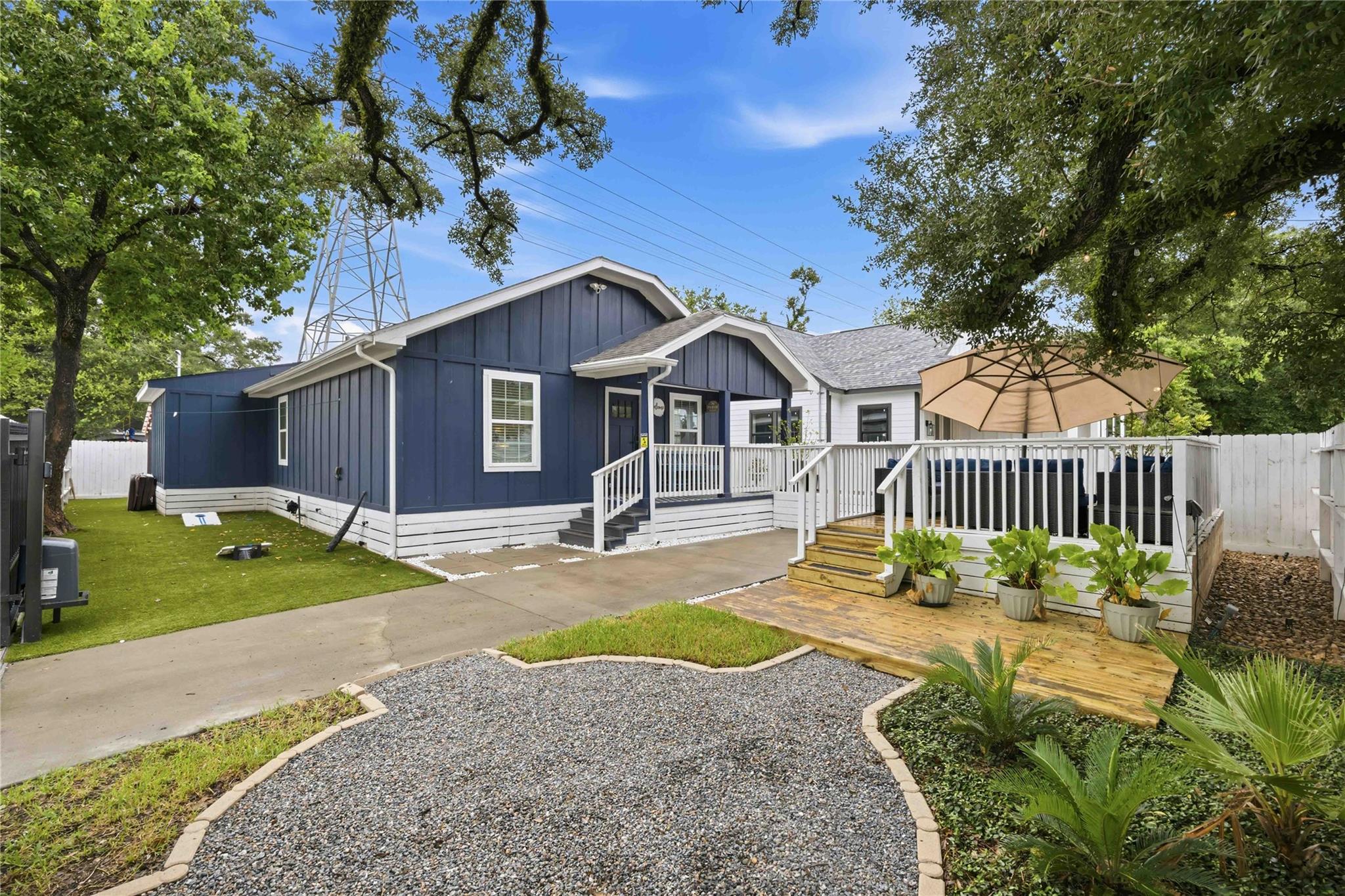 Blue-and-white house with a raised deck, tan umbrella, and gravel path through a landscaped yard.