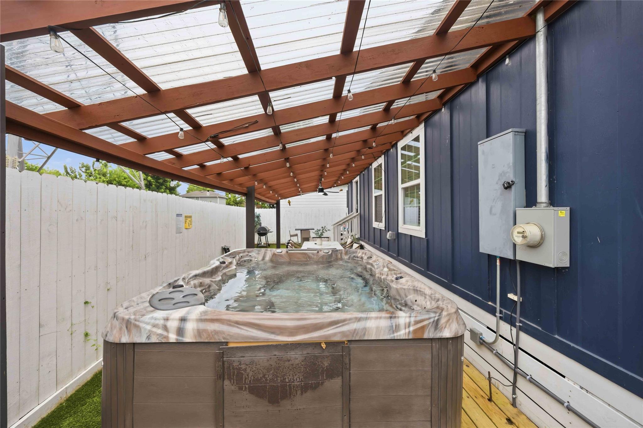 Backyard hot tub beneath a wood pergola with string lights, beside a navy blue house.