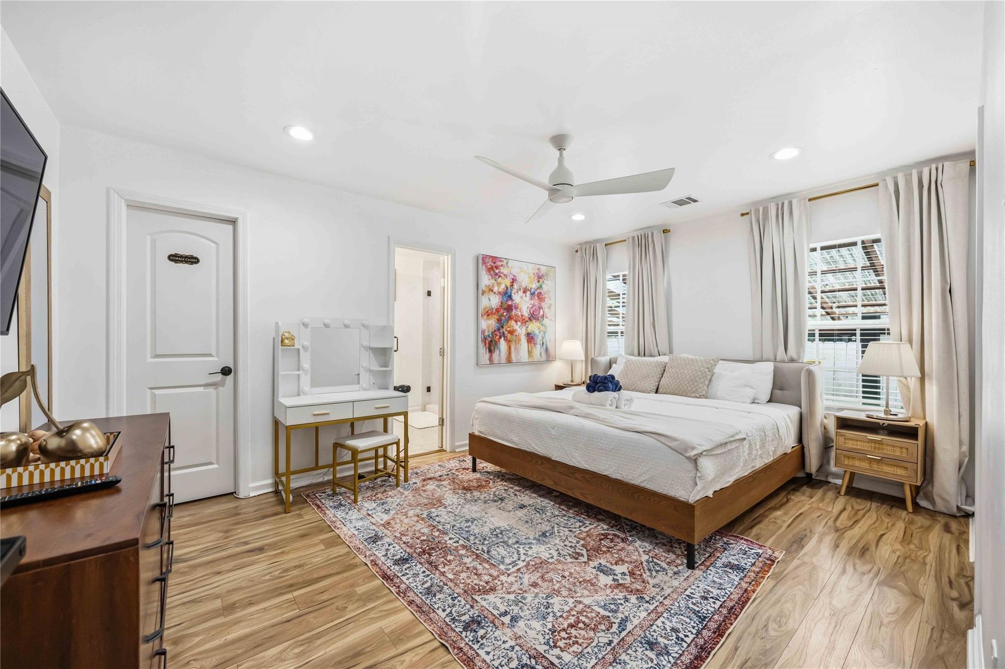 Bright bedroom with a white-dressed wooden bed, patterned rug, and curtained windows.