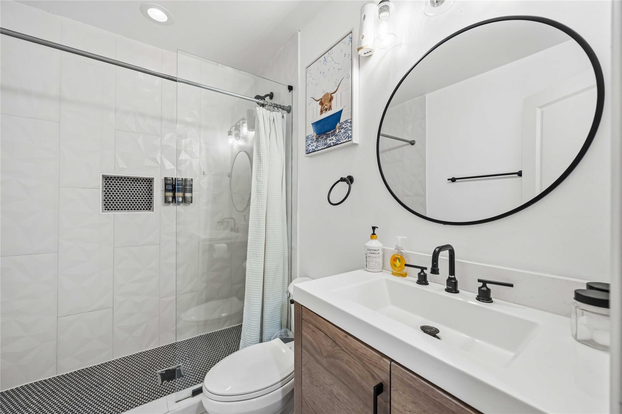 Bathroom with a glass shower, wood vanity, and large round black-framed mirror.