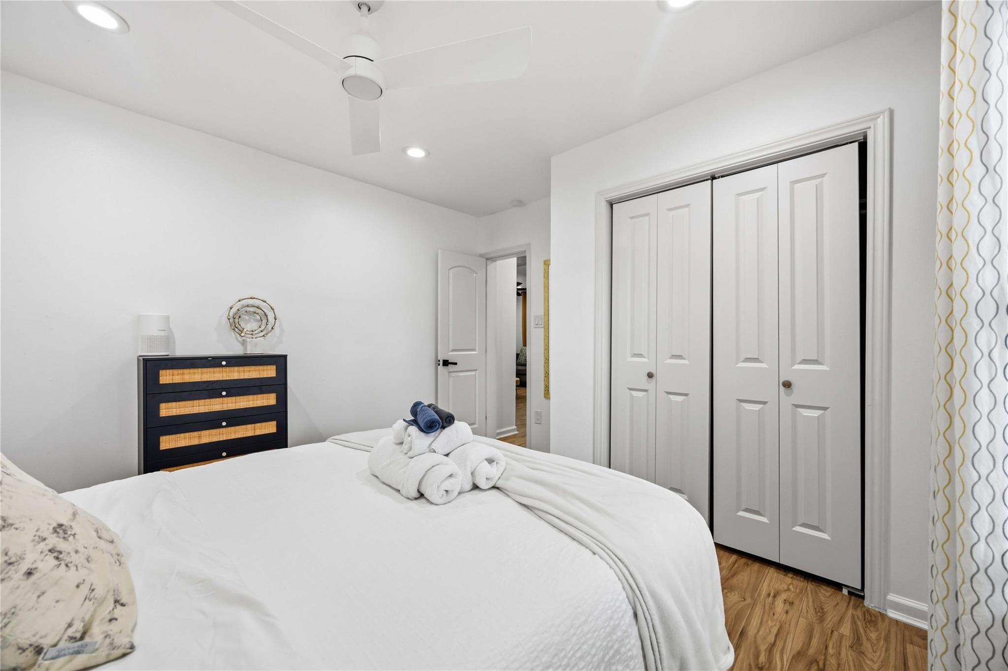Staged bedroom with a white bed, folded towels, woven-front dresser, and white closet doors.