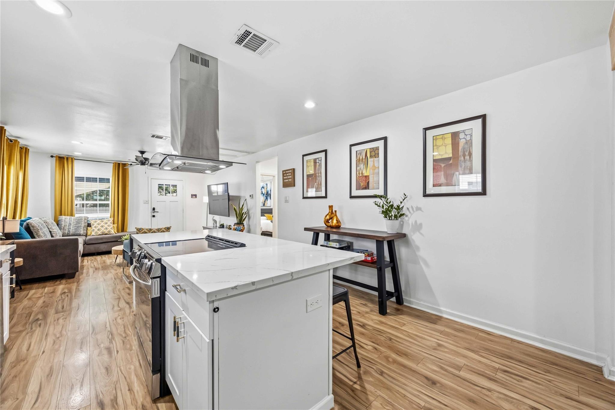 Open-plan kitchen with a large white island, stainless-steel range hood, wood-look floors, and mustard-yellow curtains.