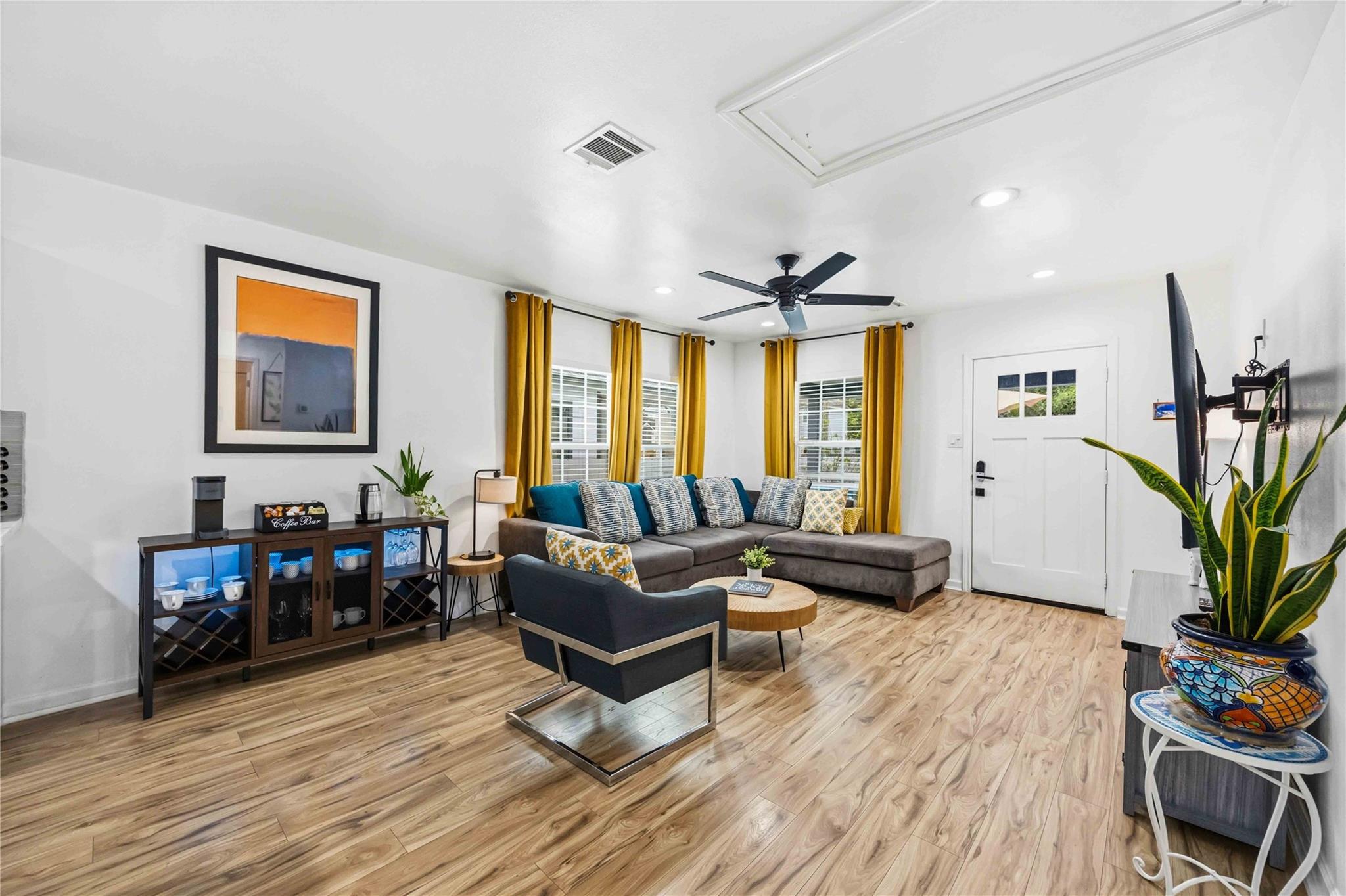 Living room with gray sectional, mustard curtains, wood-look floors, and a coffee station.