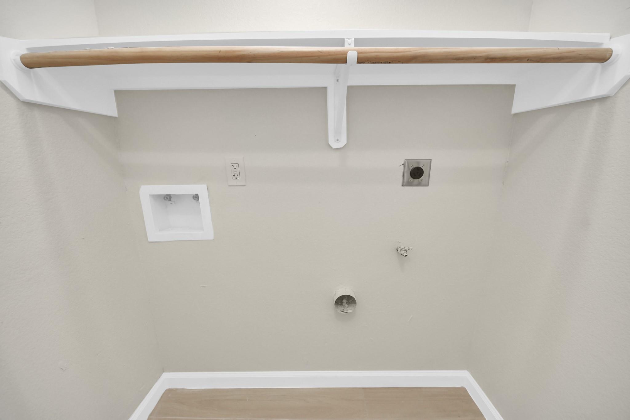 Laundry hookups beneath a white shelf and wooden hanging rod.