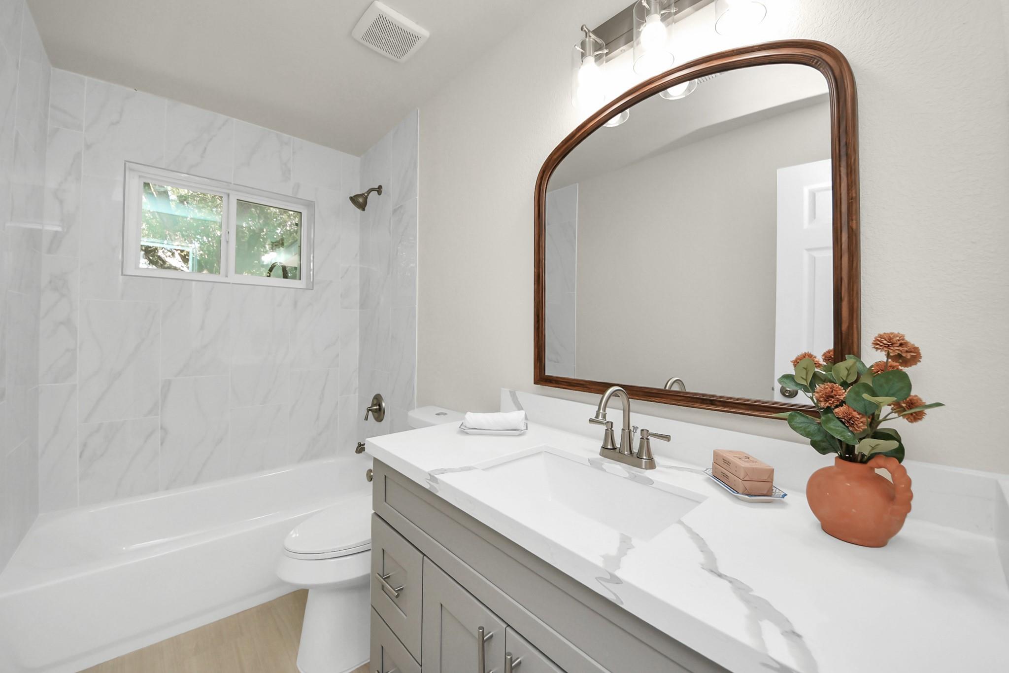 Bathroom with marble-look tub surround, gray vanity, and large arched wood-framed mirror.
