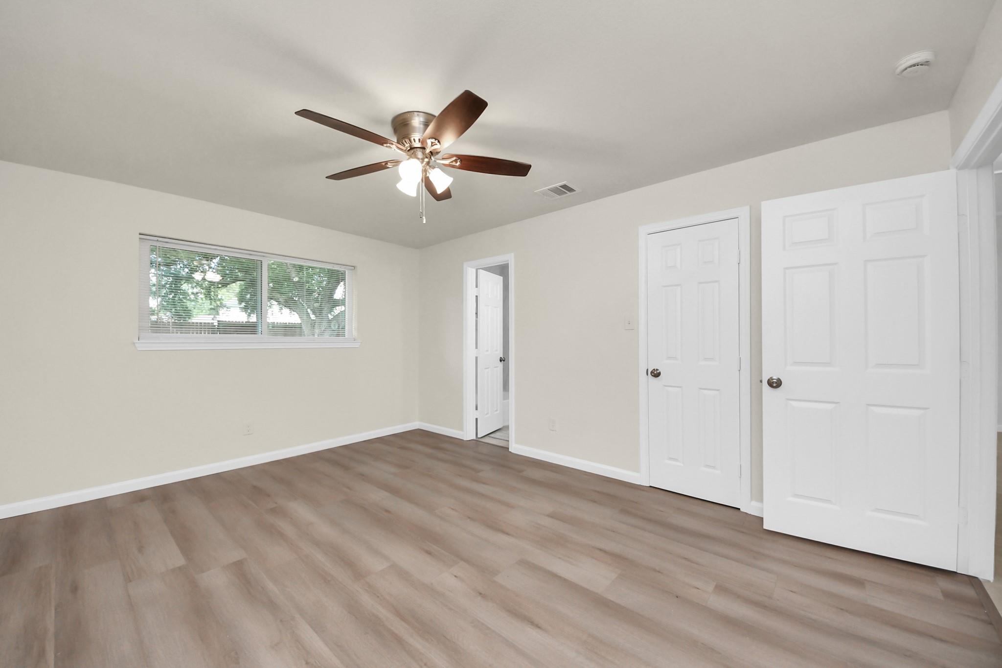 Empty bedroom with wood-look flooring, a ceiling fan, wide window, and two white closet doors.